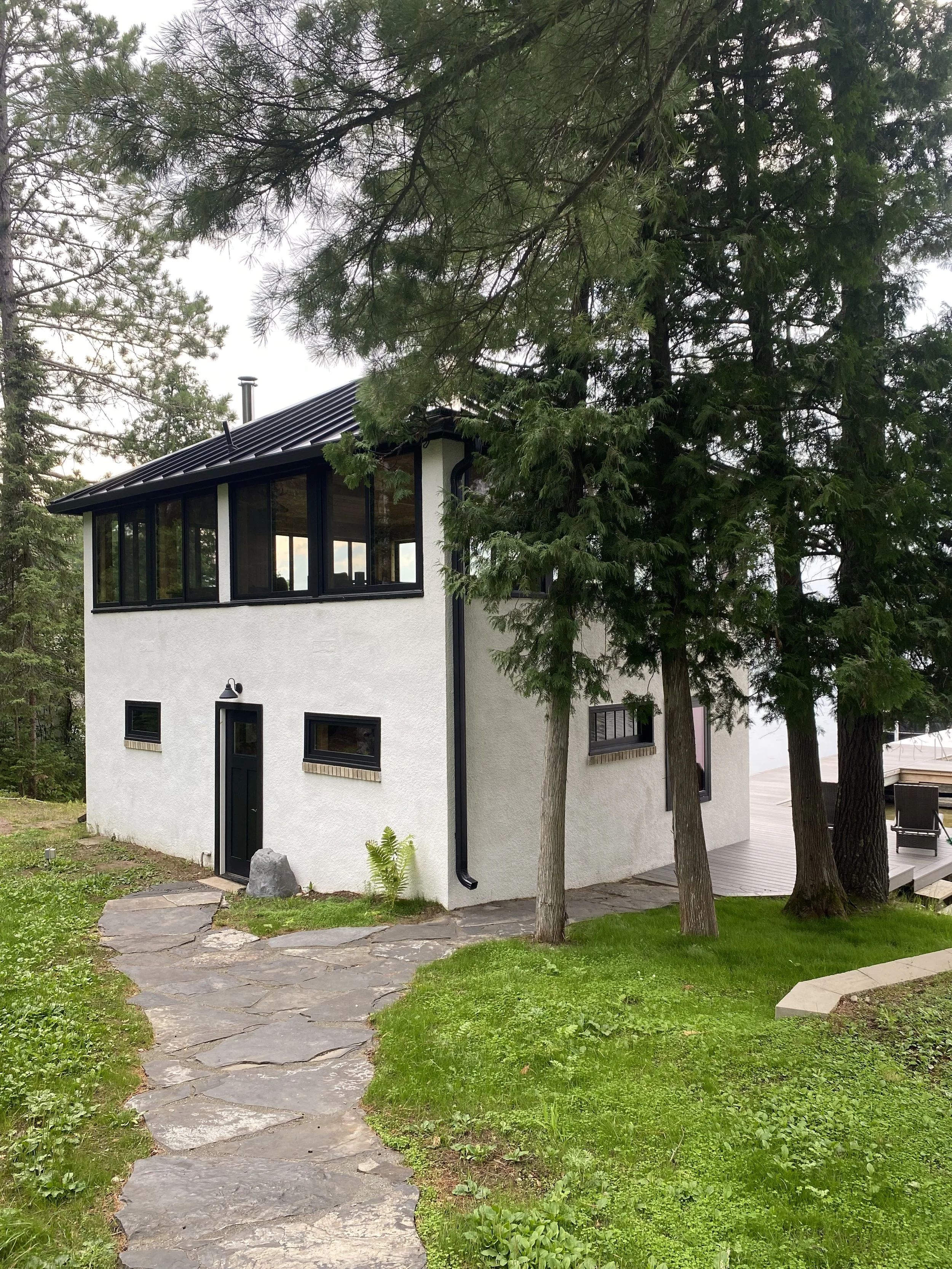 A modern white house with black-framed windows and roof, surrounded by tall pine trees and a stone pathway leading to the entrance, with clean windows