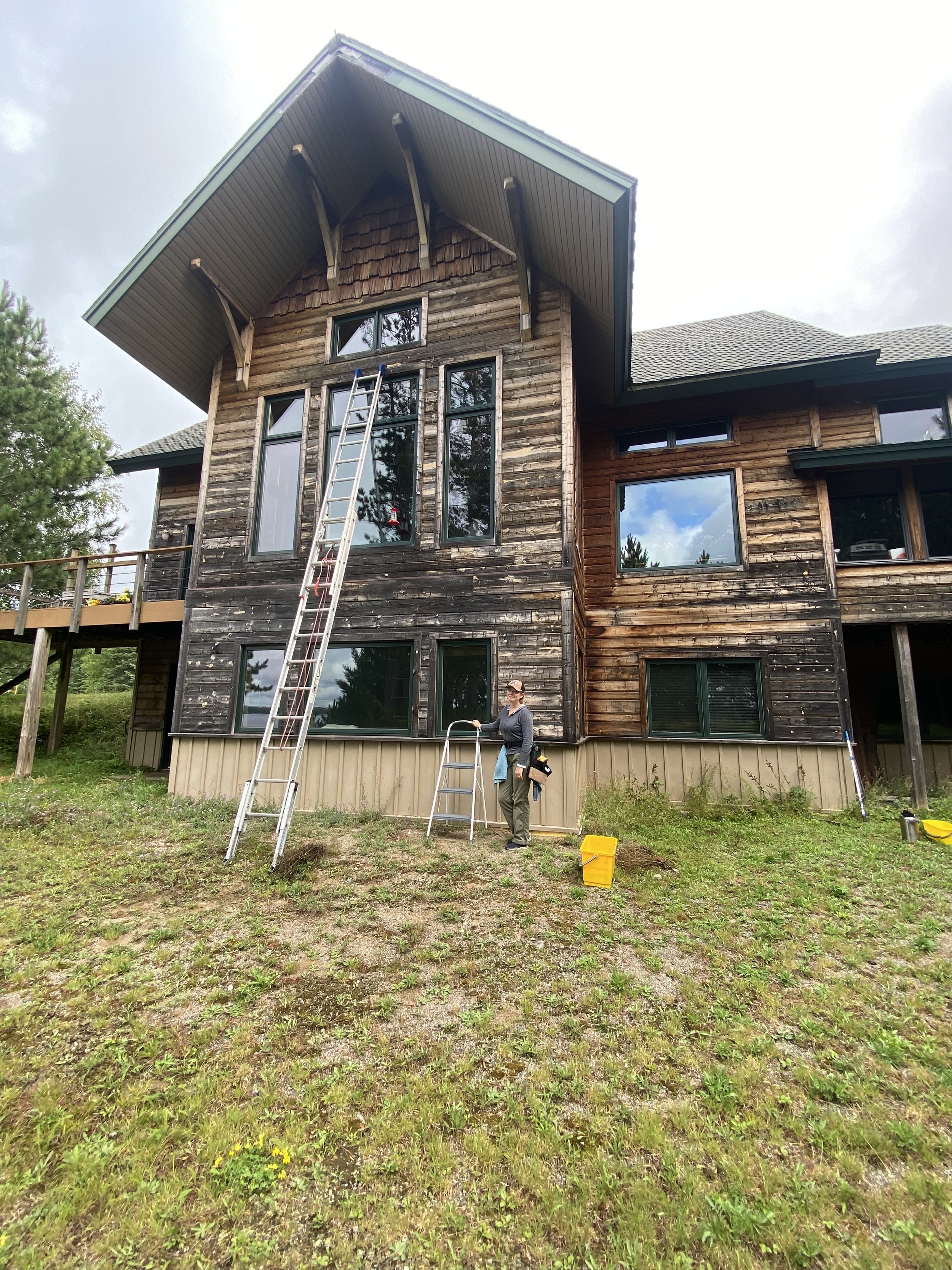 A two-story house with a wood exterior and multiple large windows, under a cloudy sky. A window cleaner is posing next to a stool she uses to get a detailed job done cleaning the windows, a tall ladder shows that the company can reach tall windows