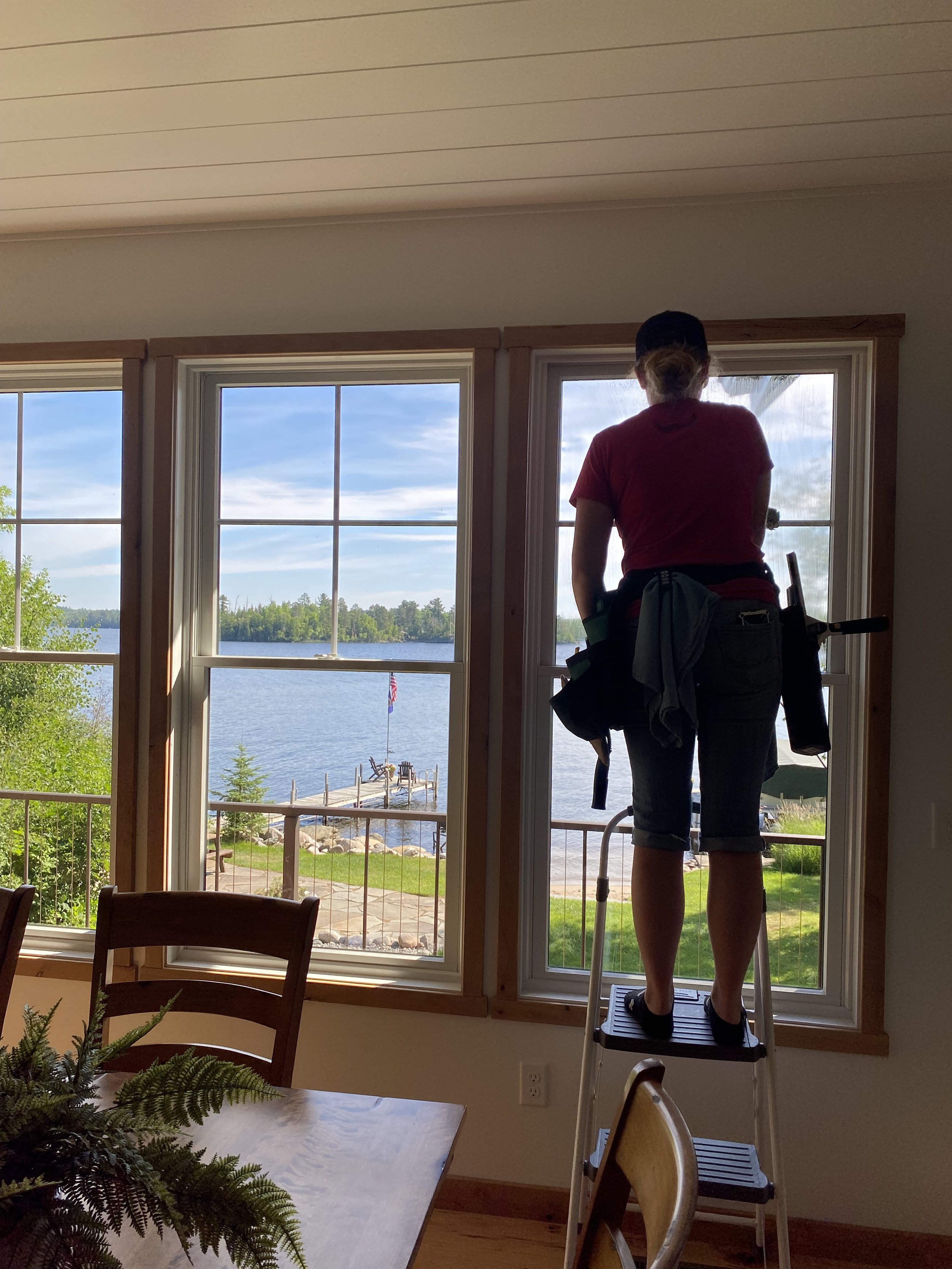 A woman standing on a step stool cleaning a large window in a room with a view of a lake and trees outside.