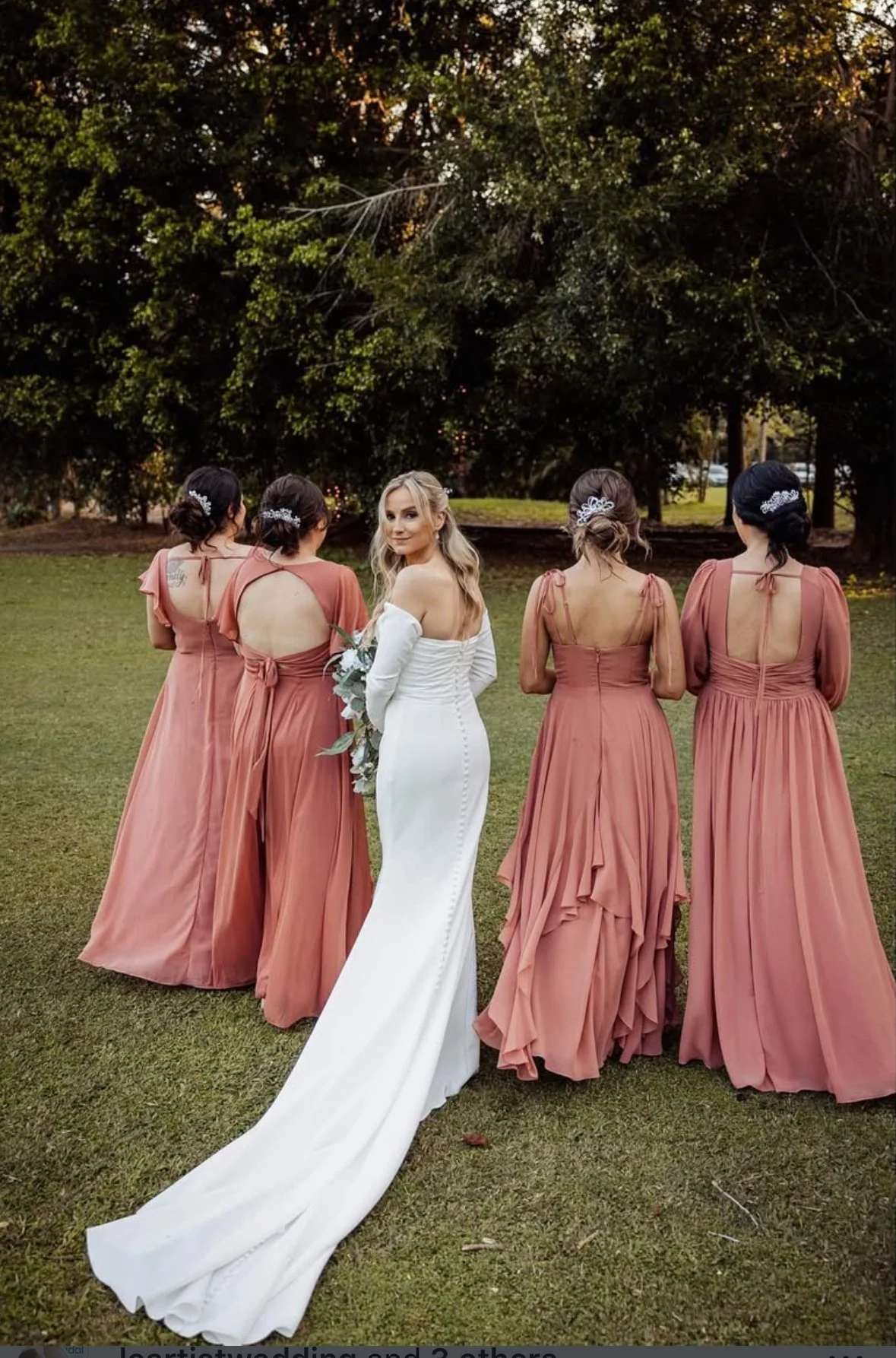 A bride in a white wedding dress holding a bouquet stands outdoors with four bridesmaids wearing matching peach-colored dresses, in a park with trees in the background.