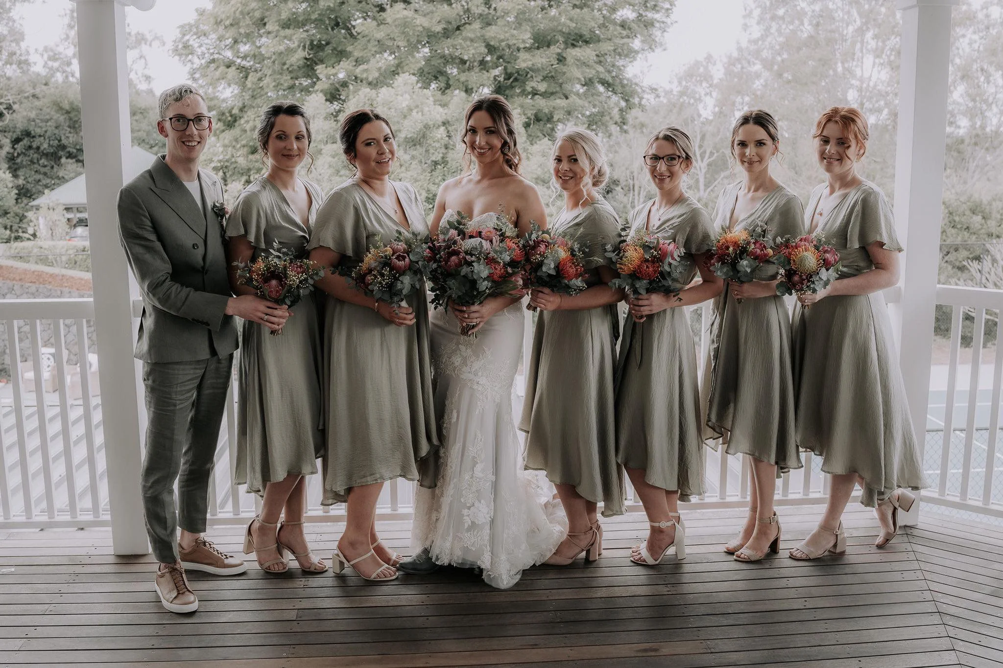 A group of eight people, including a bride in a white wedding dress, standing on a porch with a wooden floor and white railings, holding bouquets, with trees and a cloudy sky in the background.