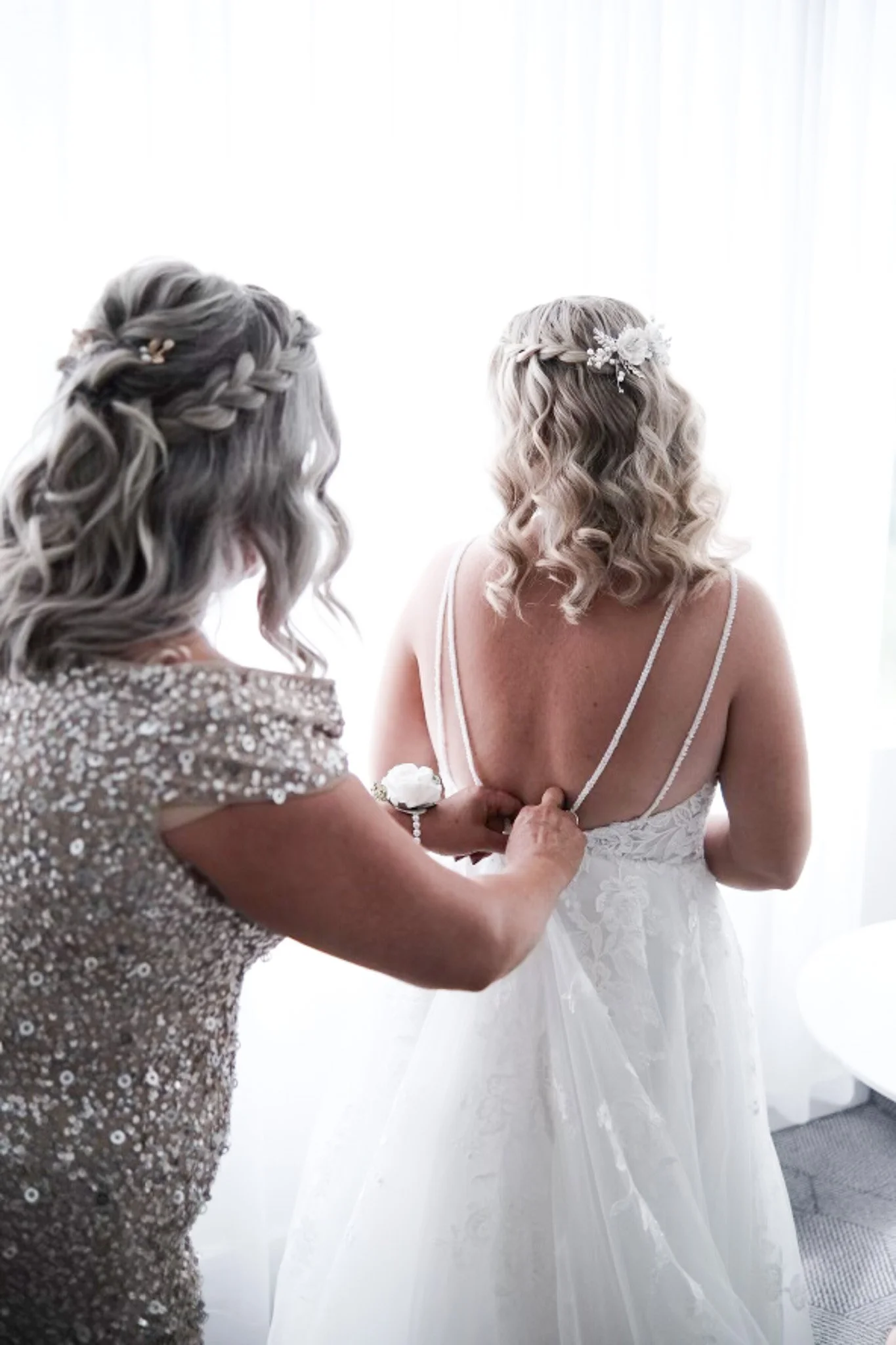 A bride in a white wedding dress is being helped by a woman in a sequin dress, possibly adjusting her dress or jewelry. The bride has curly blonde hair with a floral hairpiece. They are indoors with a bright background.