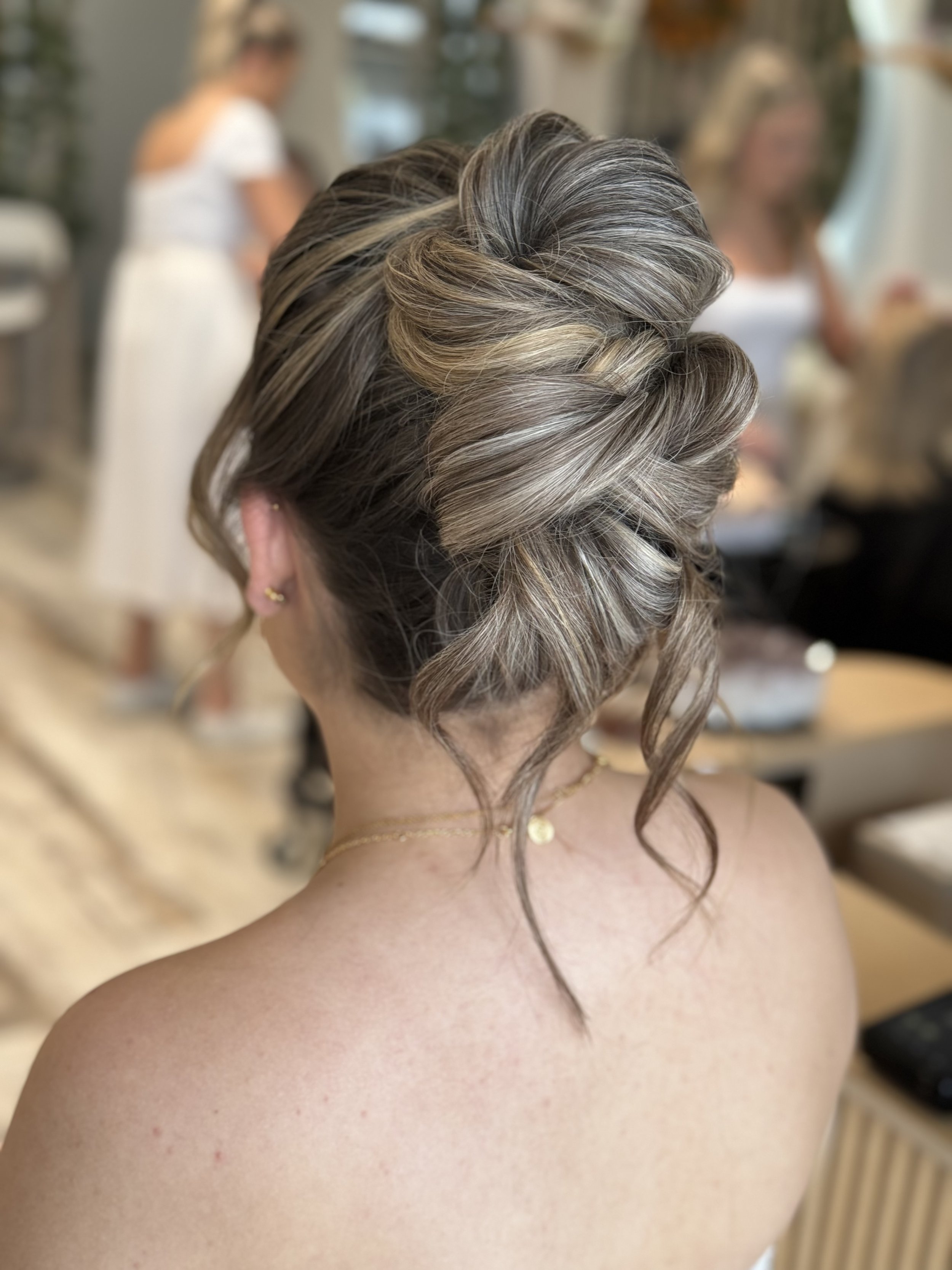 A woman with styled, curled hair in shades of gray, blonde, and brown, with a focus on the back of her head, in a salon setting. Other blurred women are visible in the background.