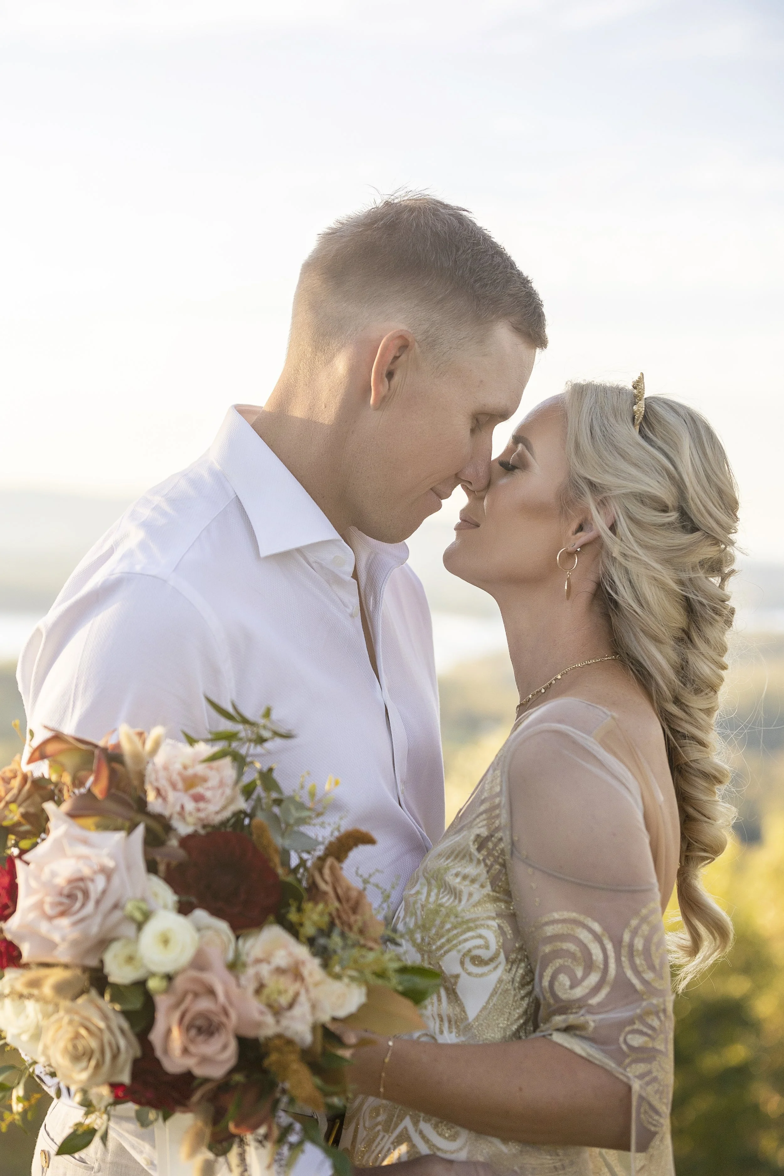 A couple in wedding attire embracing with foreheads touching outdoors during golden hour, holding a bouquet of pink, white, and burgundy flowers.