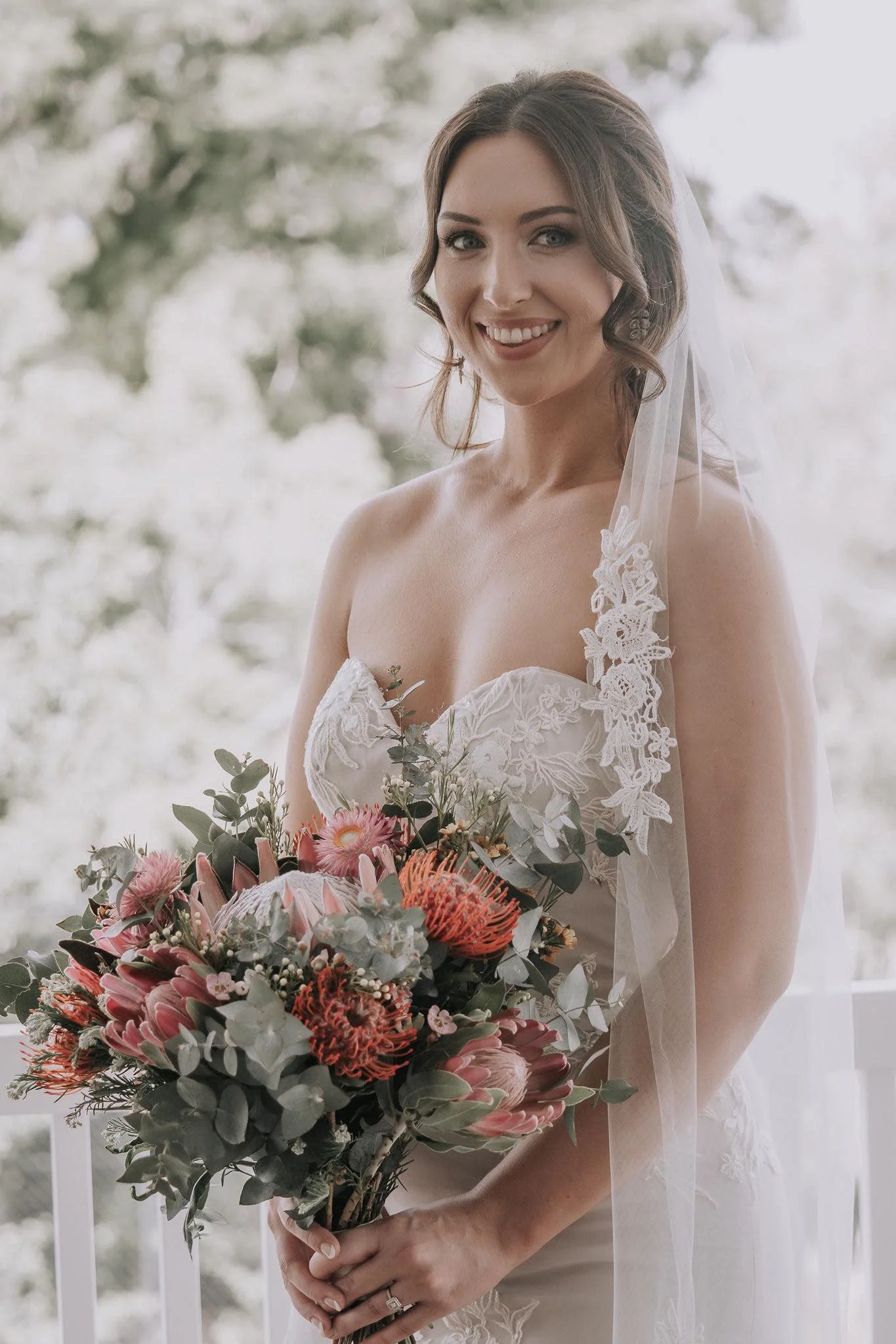 A smiling bride holding a large bouquet of pink and red flowers with green foliage, standing near a window with soft natural light.