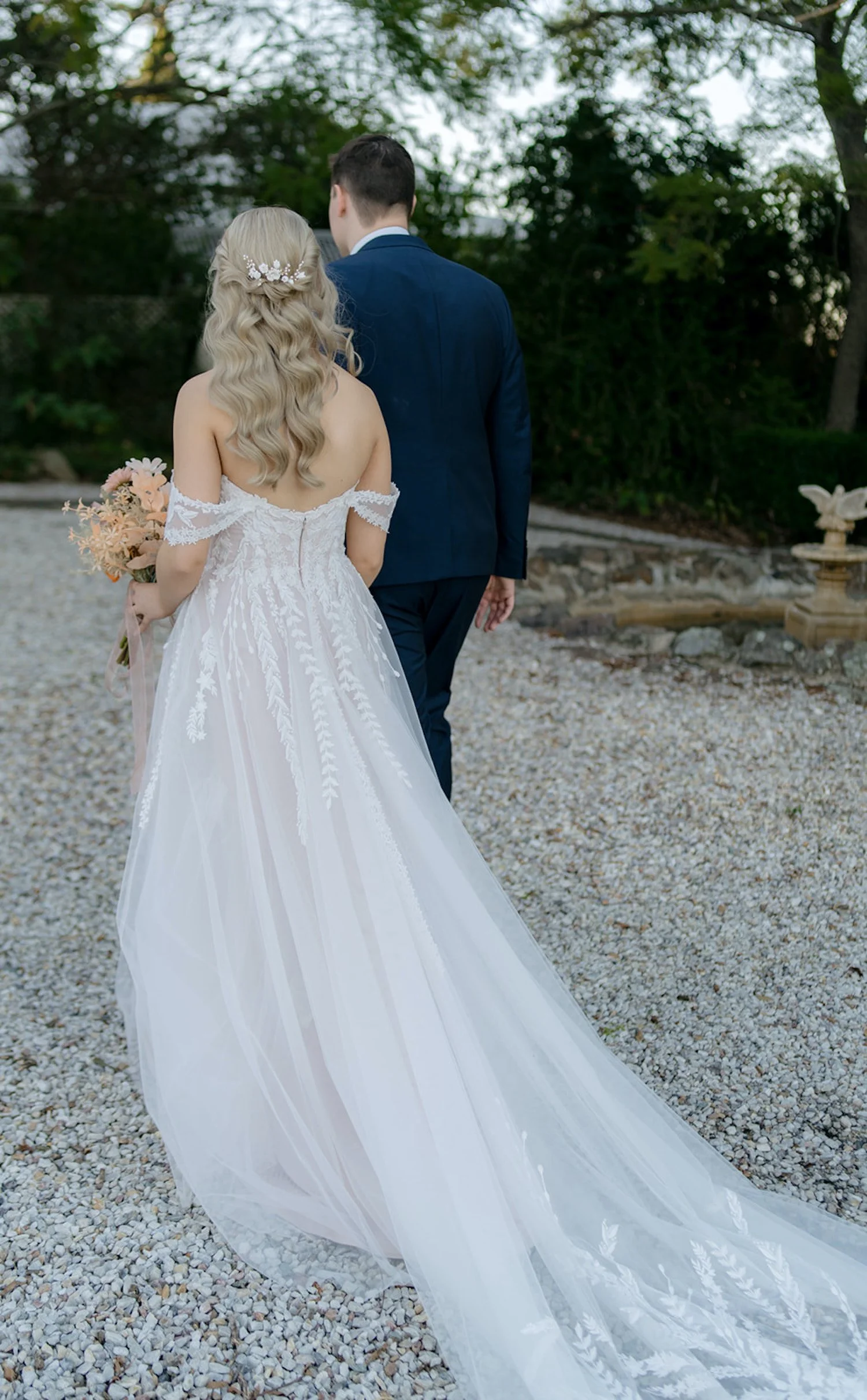 A bride and groom walking outdoors, the bride in a white lace off-the-shoulder wedding gown holding a bouquet, and the groom in a navy blue suit, with a backdrop of trees and a stone-paved area.