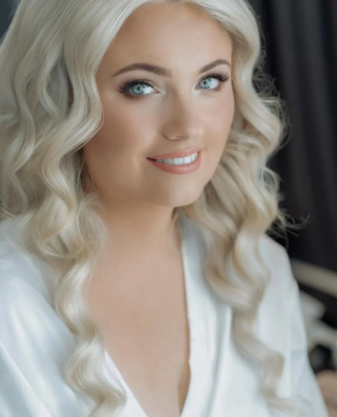 Close-up photo of a woman with long, wavy blonde hair and bright blue eyes, smiling softly, wearing a white top.