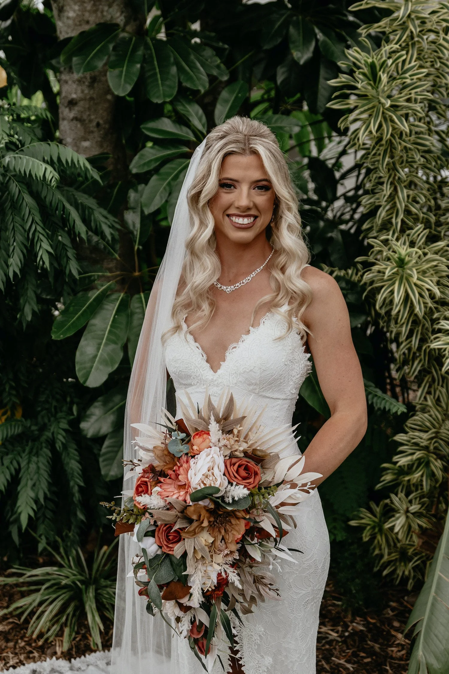 A smiling bride with long blonde hair styled in loose waves, wearing a white lace wedding dress with thin straps and a V-neckline, accessorized with a pearl necklace, standing outdoors among lush green tropical plants. She holds a large bouquet of dried and fresh flowers in shades of rust, peach, cream, and green, and has a long, sheer veil over her shoulder.