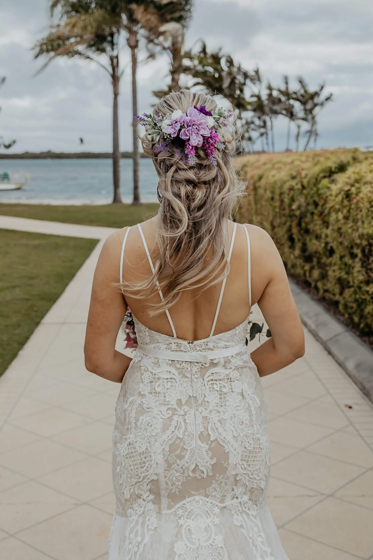 A woman wearing a white lace wedding dress with thin straps, standing outdoors with her back to the camera. She has long, wavy blonde hair adorned with a large purple and pink floral headpiece. The background features palm trees, a body of water, and cloudy sky.