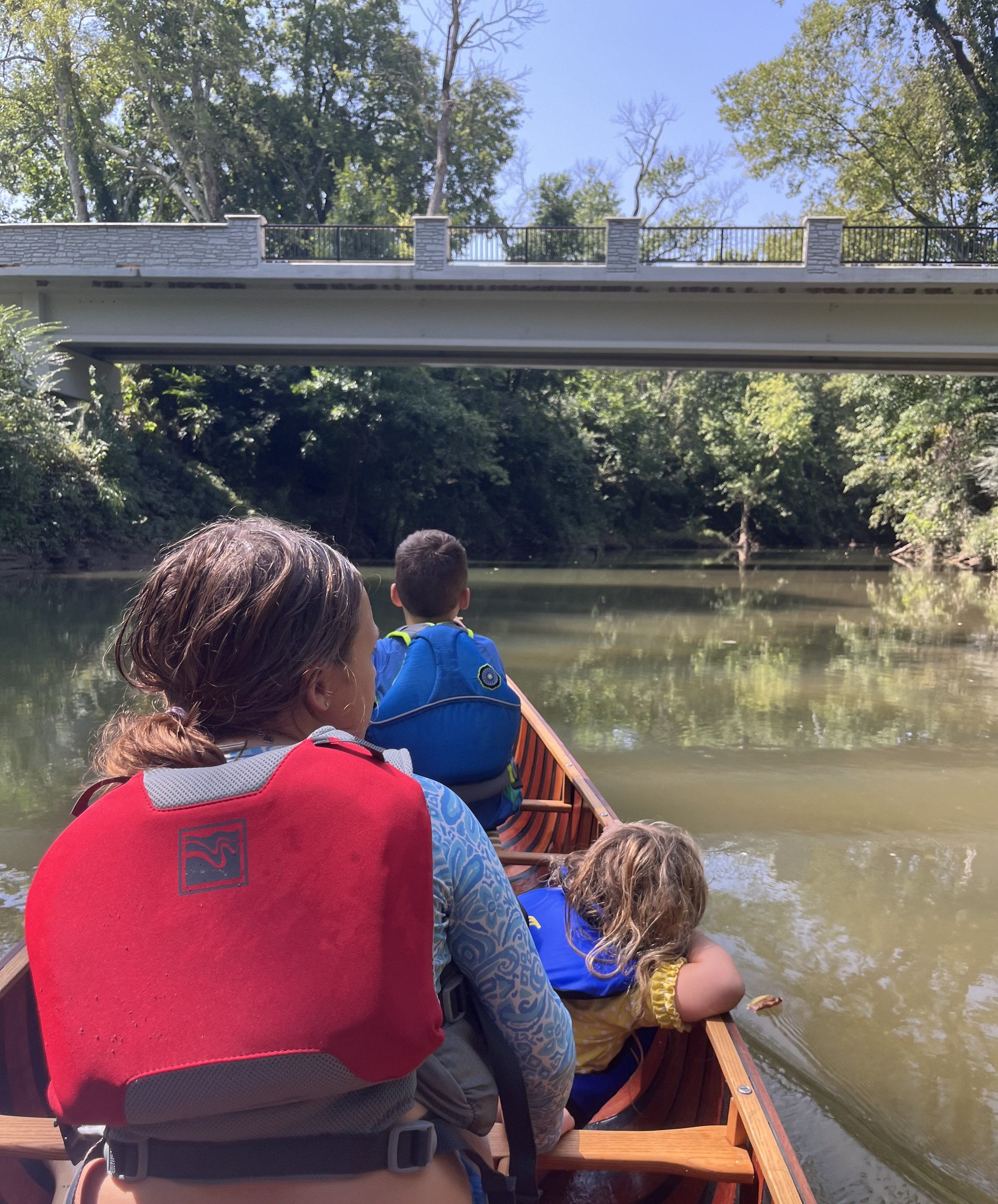People sitting in a wooden boat on a river, with a bridge overhead and lush green trees along the riverbank. One person is reaching into the water.