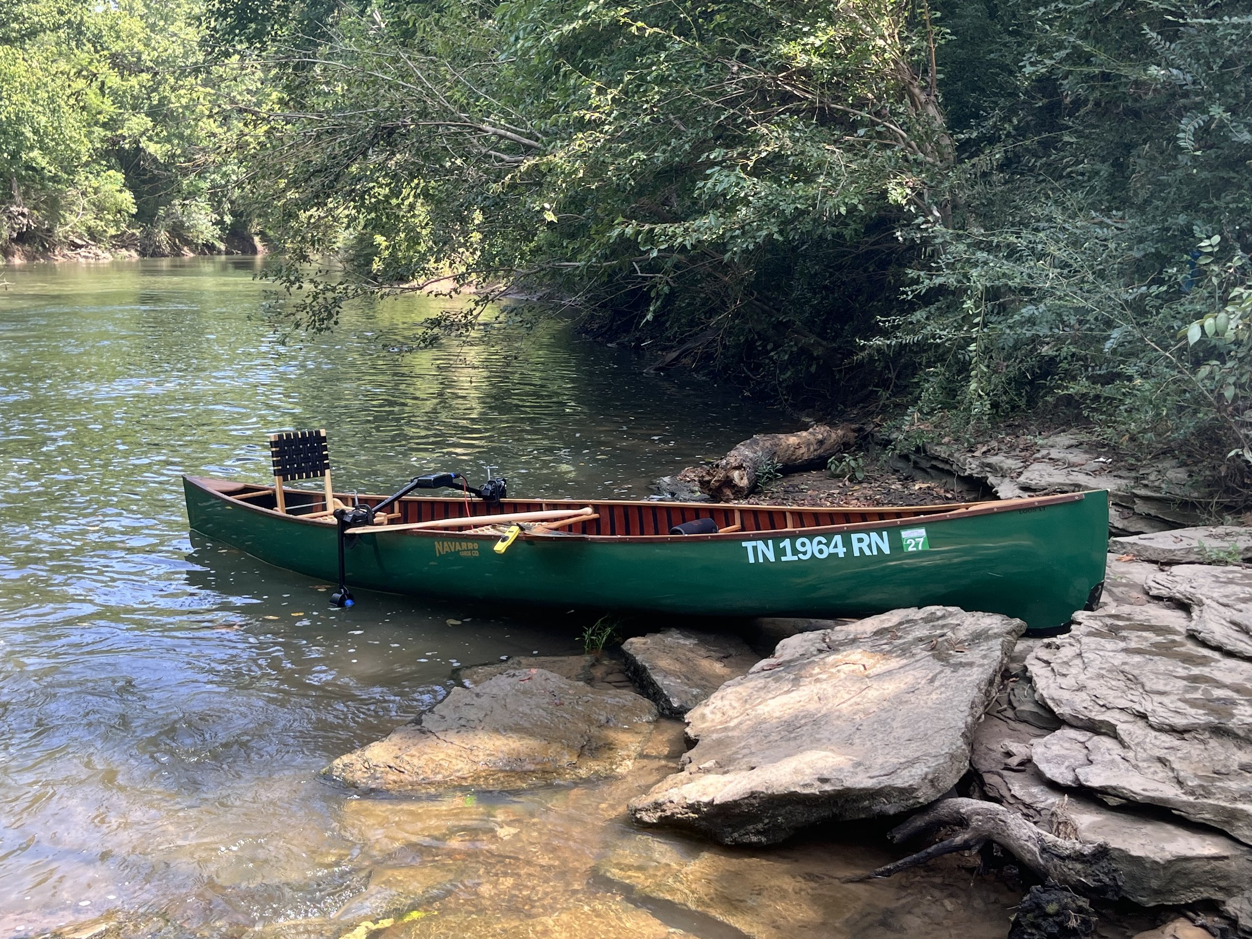 A green canoe docked by a rocky shoreline on a river surrounded by dense trees with lush green foliage.