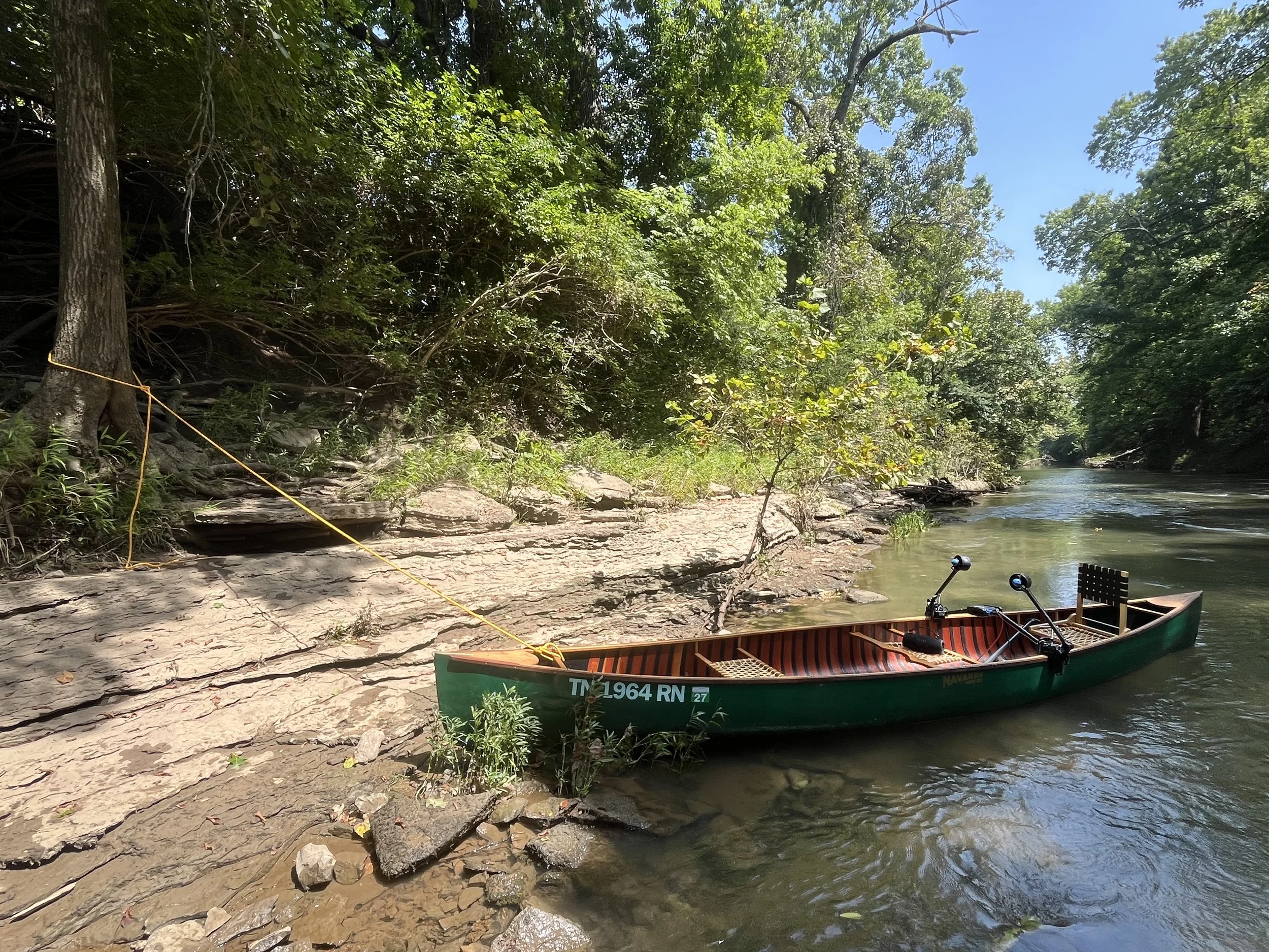 A docked boat on a riverbank surrounded by lush green trees and rocks with a clear blue sky overhead.
