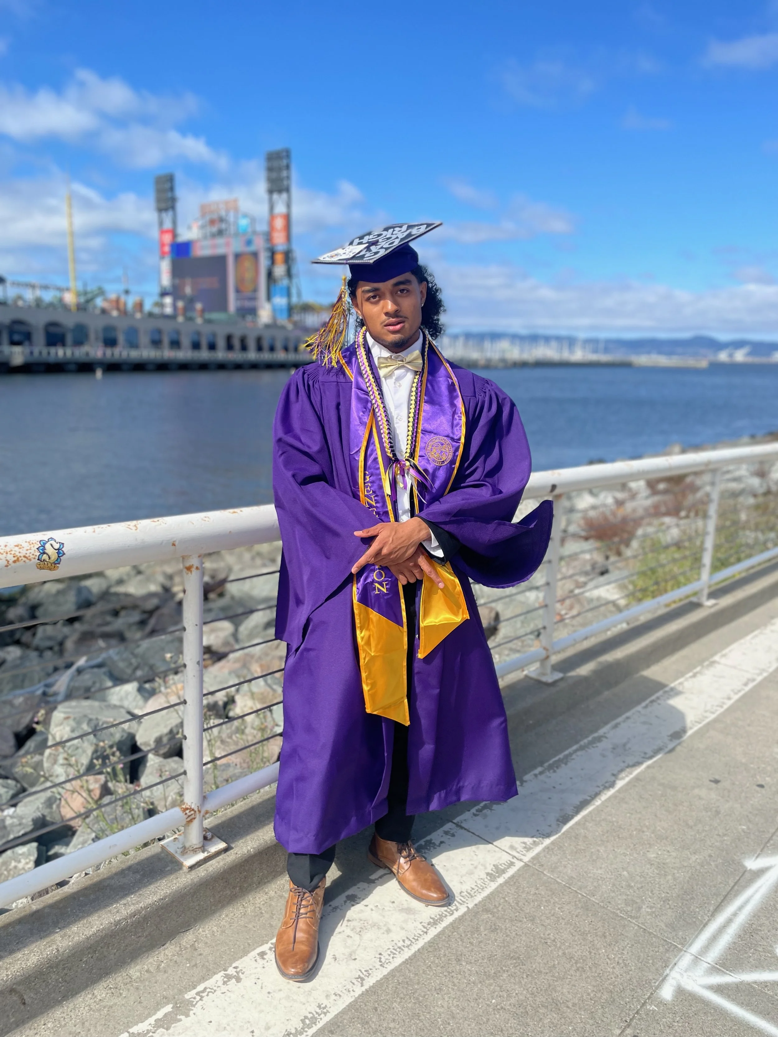 Young man in a purple graduation gown and cap with a gold tassel, standing outdoors by a waterfront with a bridge and a stadium in the background.