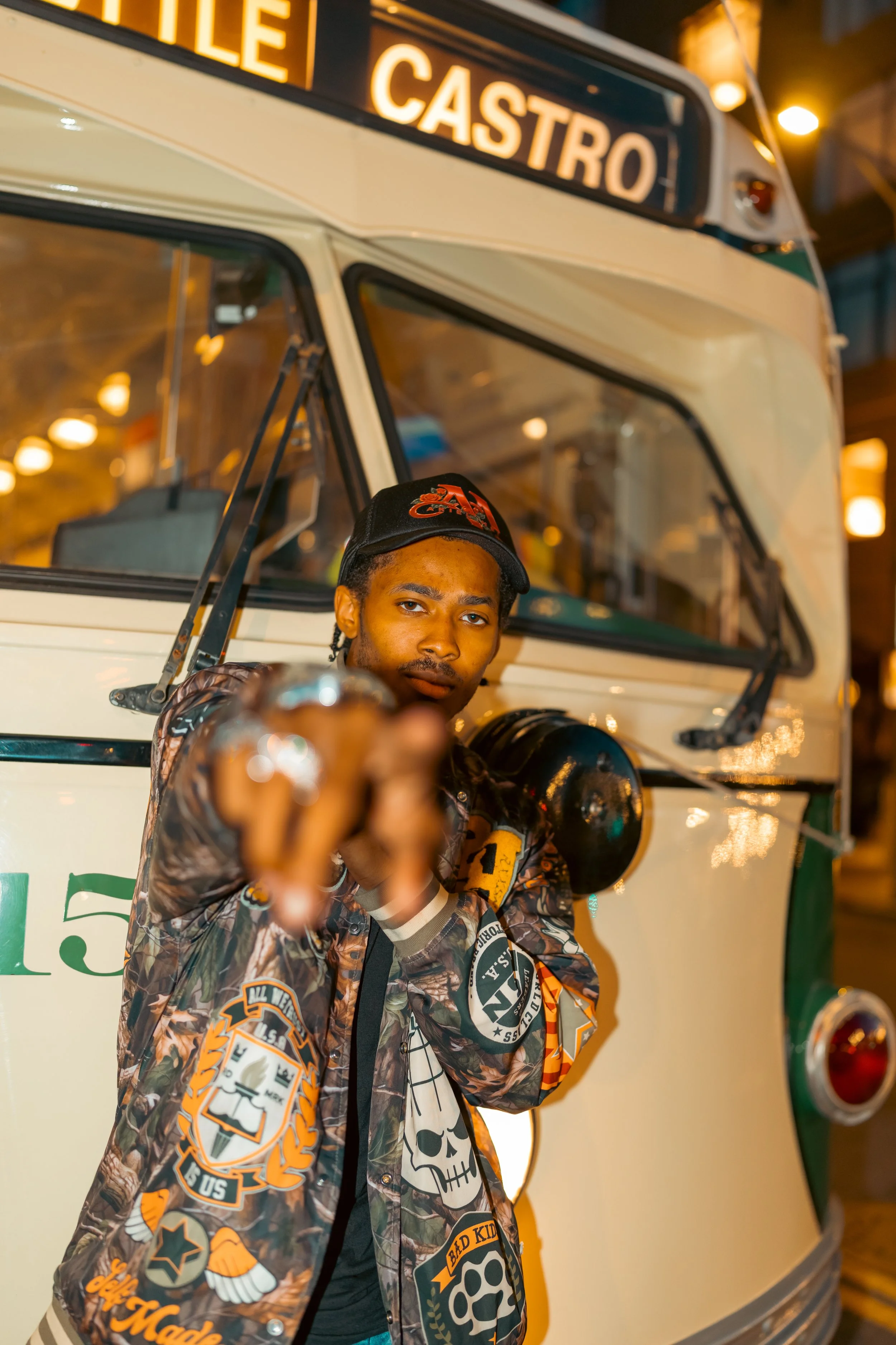 A young man with a hat and a patterned jacket pointing towards the camera, standing in front of a vehicle with the sign 'ILE CASTRO' at night.