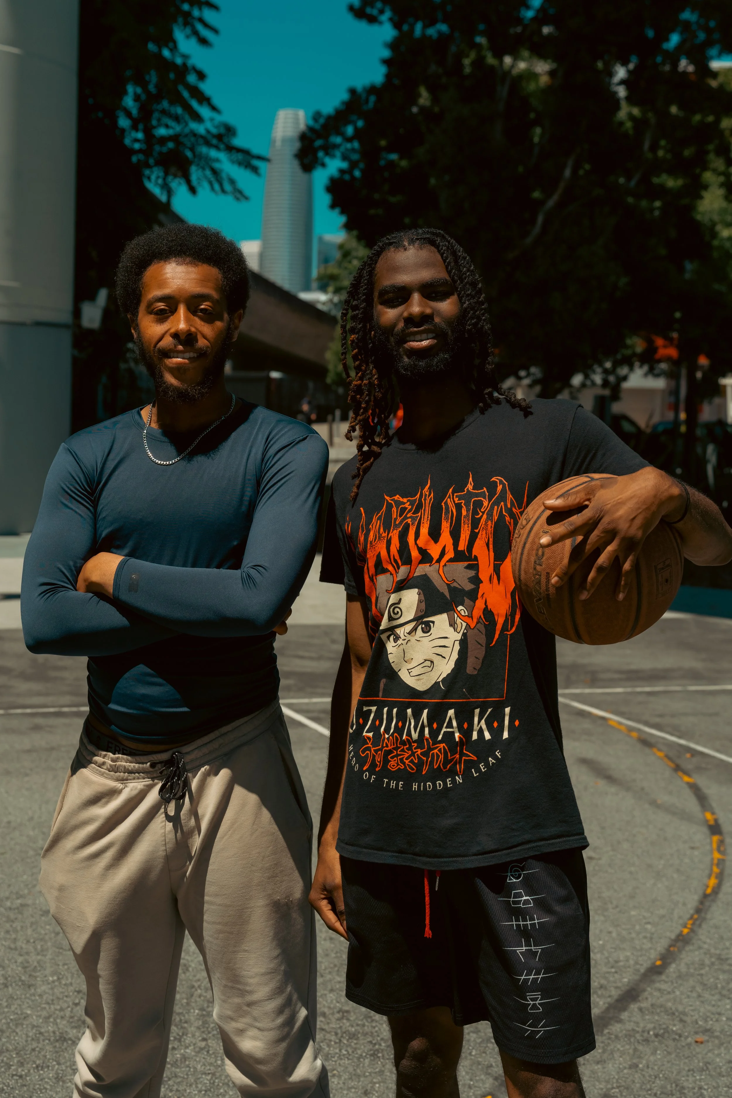Two young men standing outdoors on a sunny day, one with arms crossed and the other holding a basketball, with trees and a city skyline in the background.