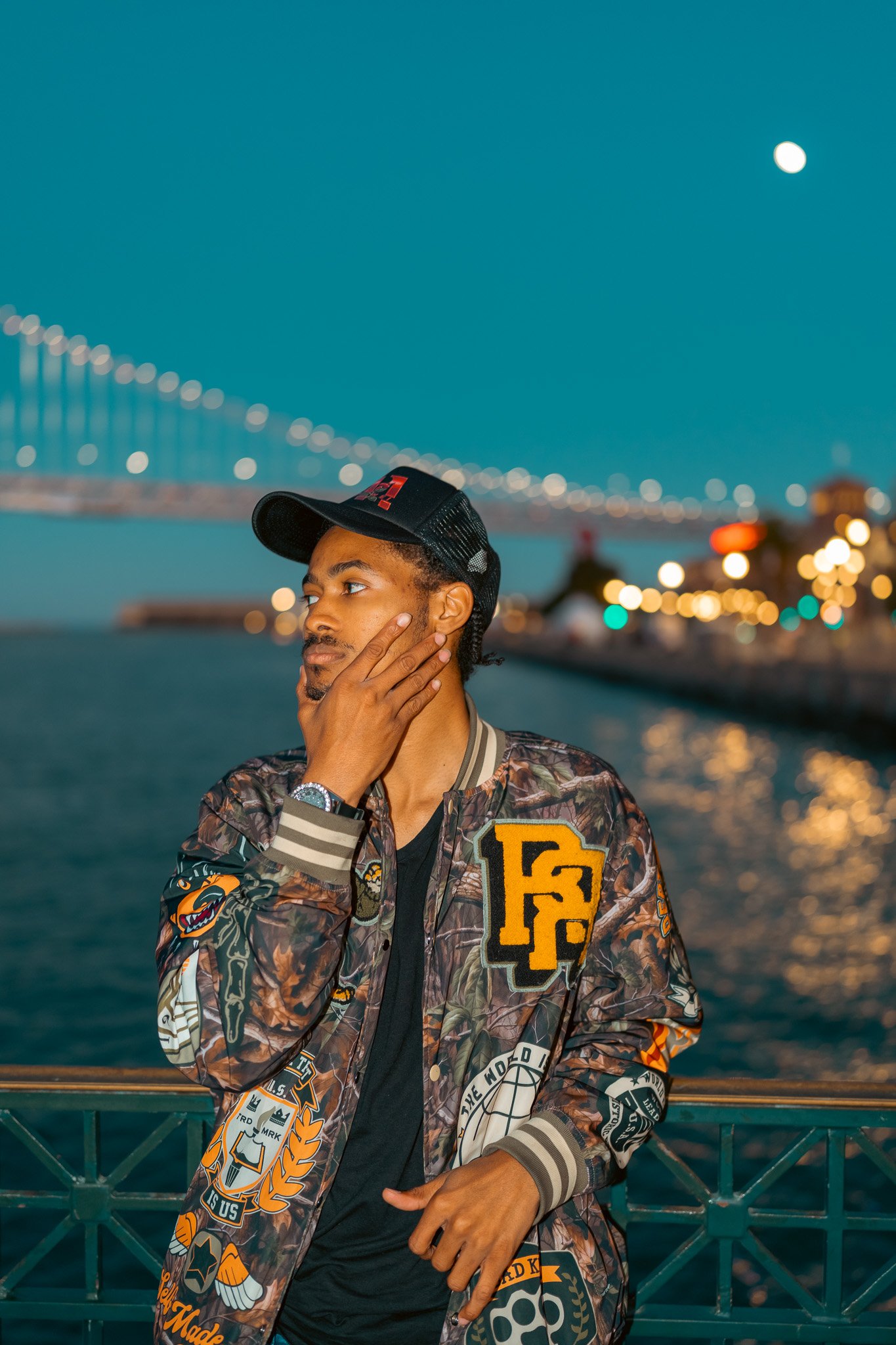 A young man with a shaved head wearing a patterned jacket and cap, standing on a bridge at dusk with city lights and the moon in the sky.
