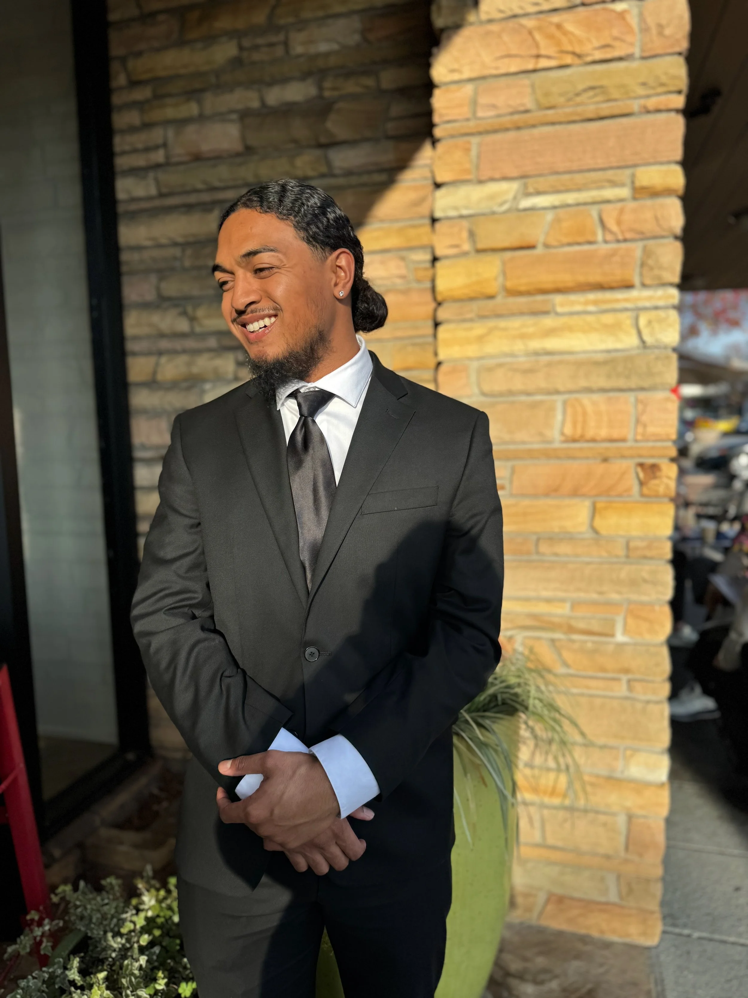 A young man in a black suit and tie standing outdoors in front of a brick wall, smiling and looking to the side.
