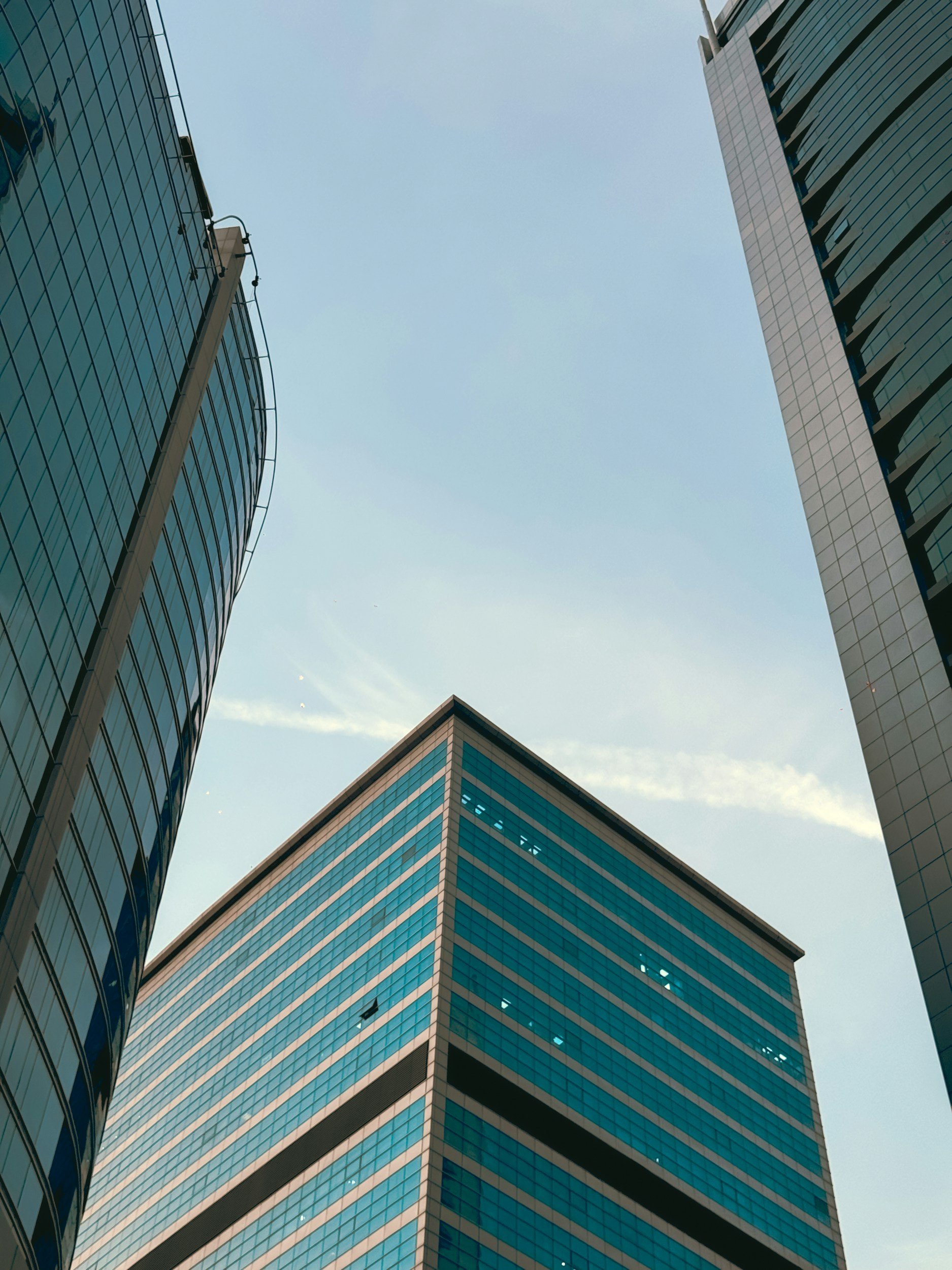 Looking up at skyscrapers with glass facades in a cityscape, showing reflections of the sky and neighboring buildings against a partly cloudy sky.