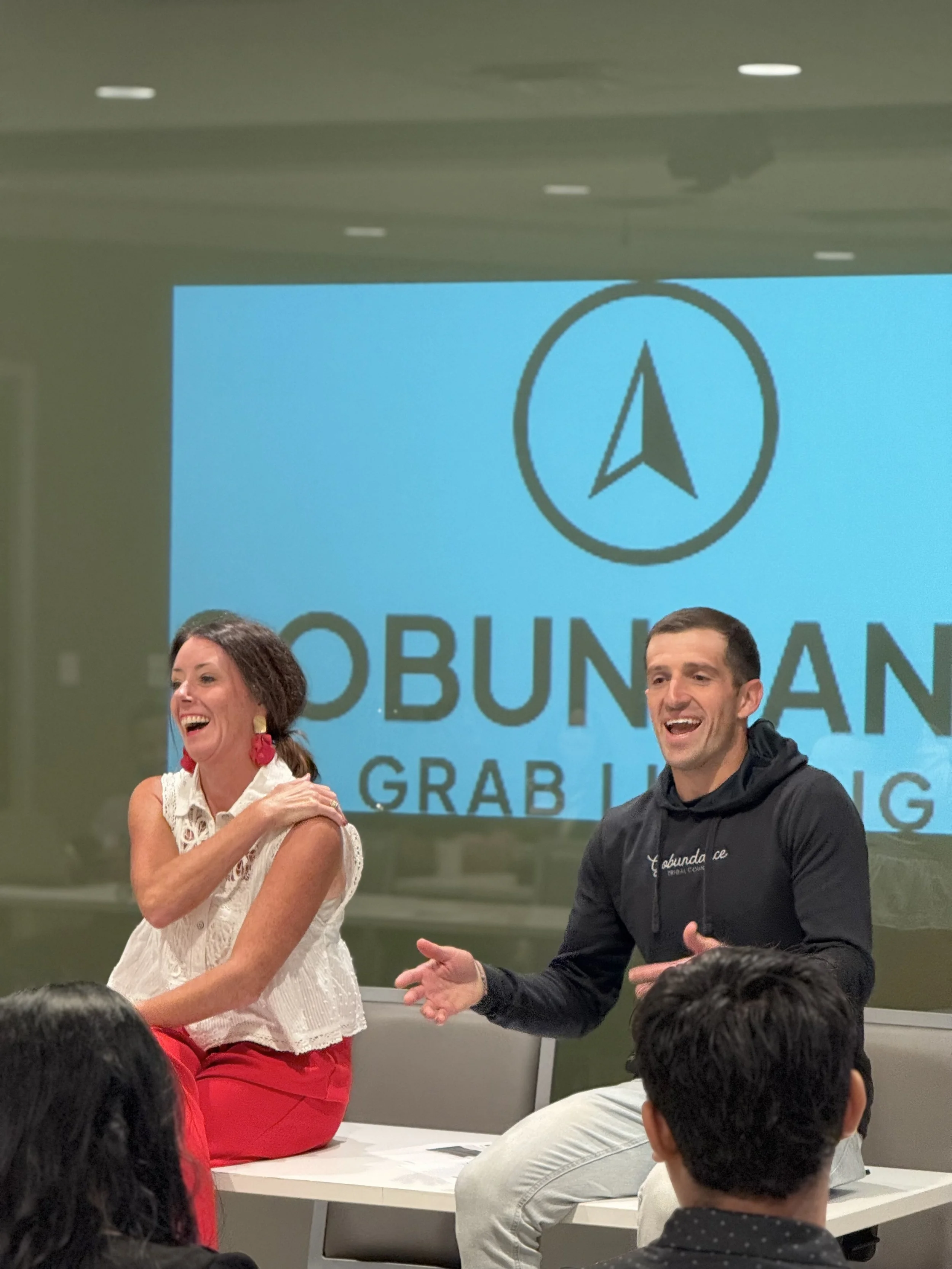 A woman and a man are standing at the front of a room, smiling and engaging with an audience. The woman has curly hair, is wearing a white sleeveless top with red pants, and is laughing. The man has short dark hair, is wearing a black jacket with white text, and is gesturing with his hands. Behind them is a large screen with a logo featuring a circle and an arrow pointing upward, along with the text 'OBUNDA.'