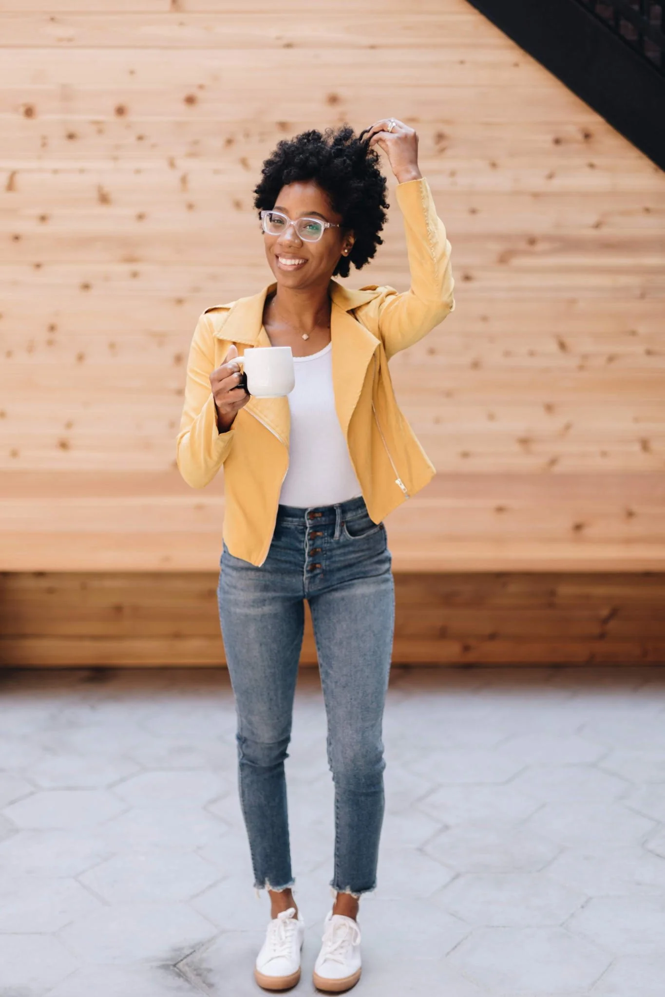 A woman with curly hair, glasses, and a bright smile holds a white mug in her right hand while adjusting her hair with her left hand. She is wearing a yellow jacket, a white top, high-waisted jeans, and white sneakers. She is standing on a cracked concrete floor against a background of polished wood paneling and a black railing.