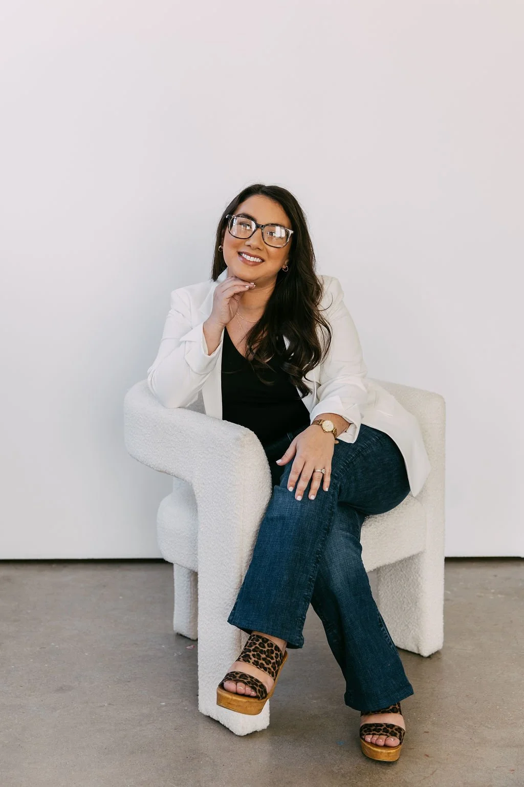 A woman with long dark hair, glasses, and a white blazer sitting on a textured white armchair against a plain white wall, smiling with her chin resting on her hand.