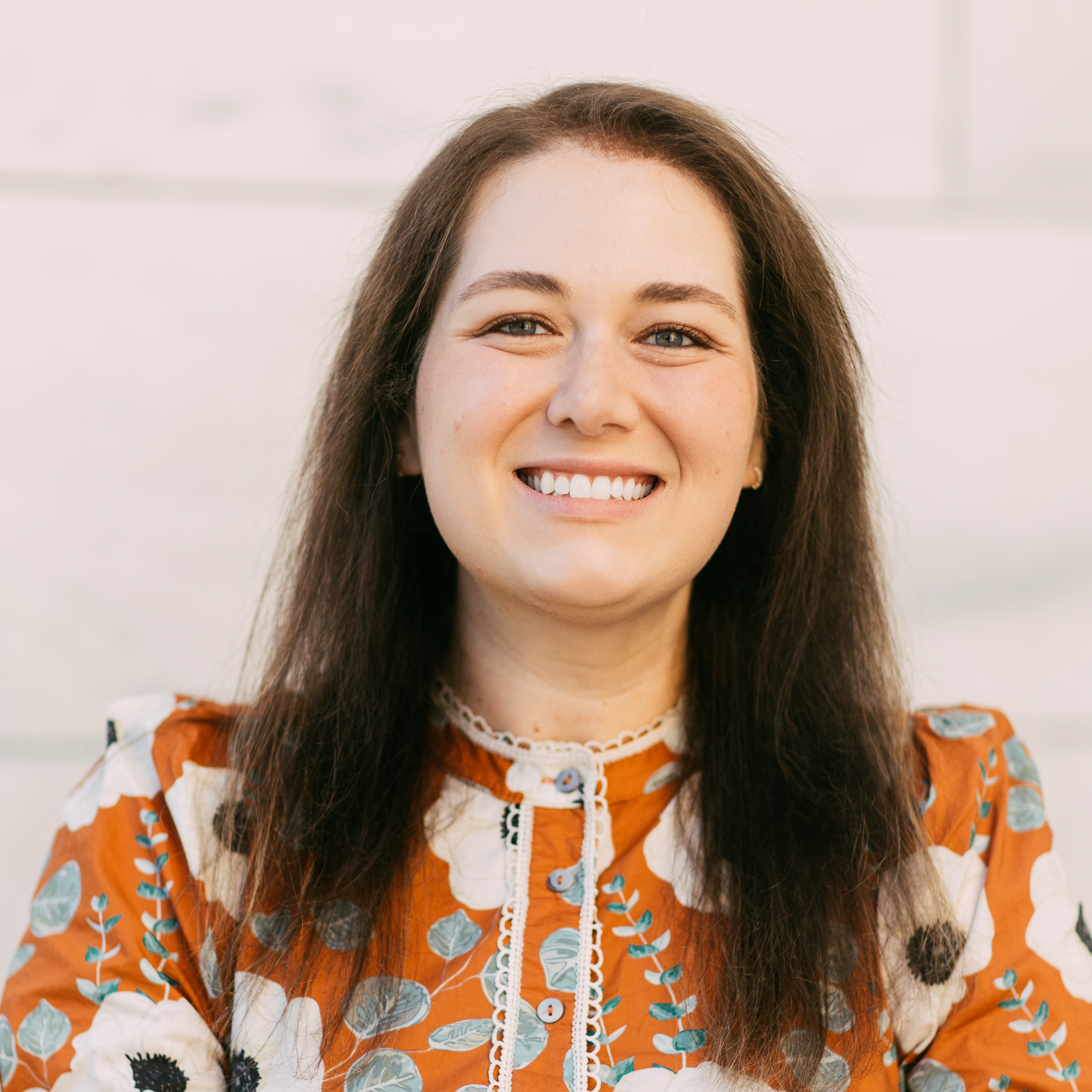 Close-up of a smiling woman with long brown hair, wearing a patterned top with orange, white, and blue floral designs, and lace detailing around the collar.