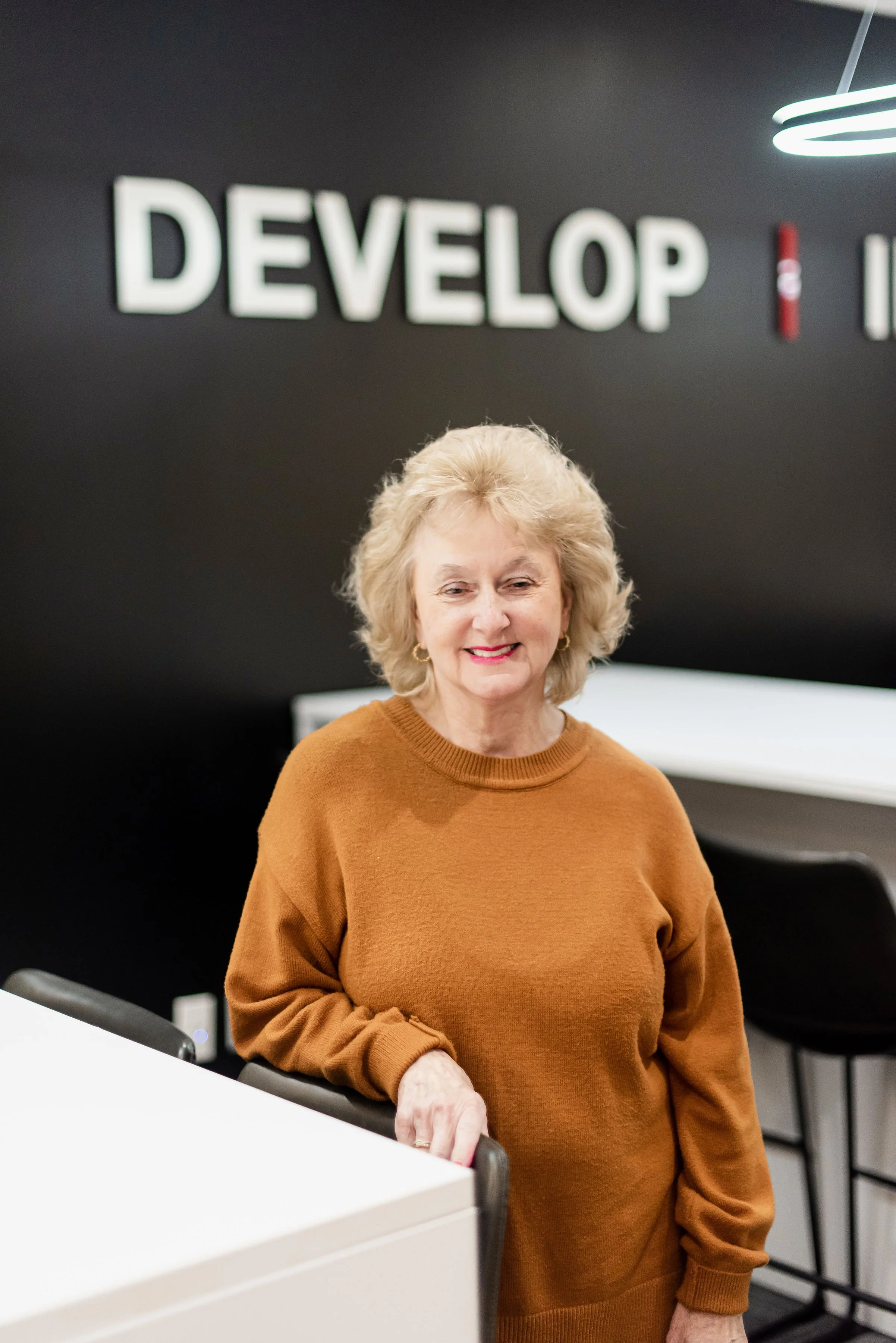 A woman with blonde hair wearing a brown sweater standing in an office or conference room. In the background, there is a black wall with large illuminated text displaying the word 'DEVELOP'.