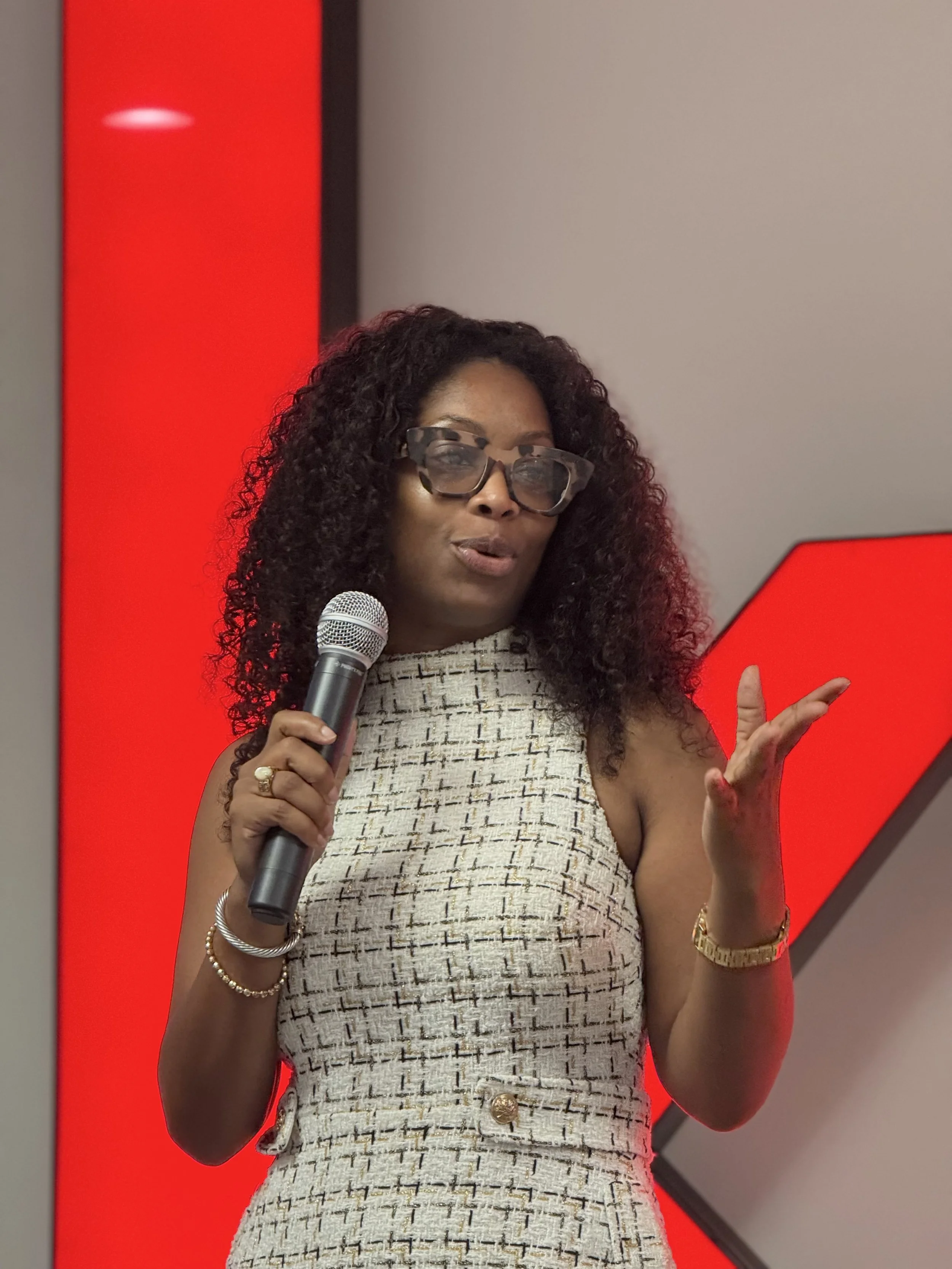 A woman with curly black hair wearing glasses, a sleeveless patterned dress, and gold jewelry holds a microphone and gestures with her left hand during a presentation or speech, standing in front of a wall with red and gray sections.