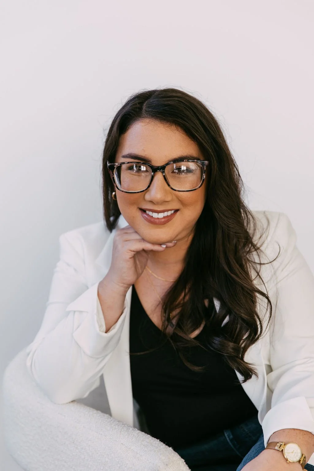 A woman with long dark hair, glasses, and a white blazer sitting on a cream-colored chair, smiling, against a plain white background.