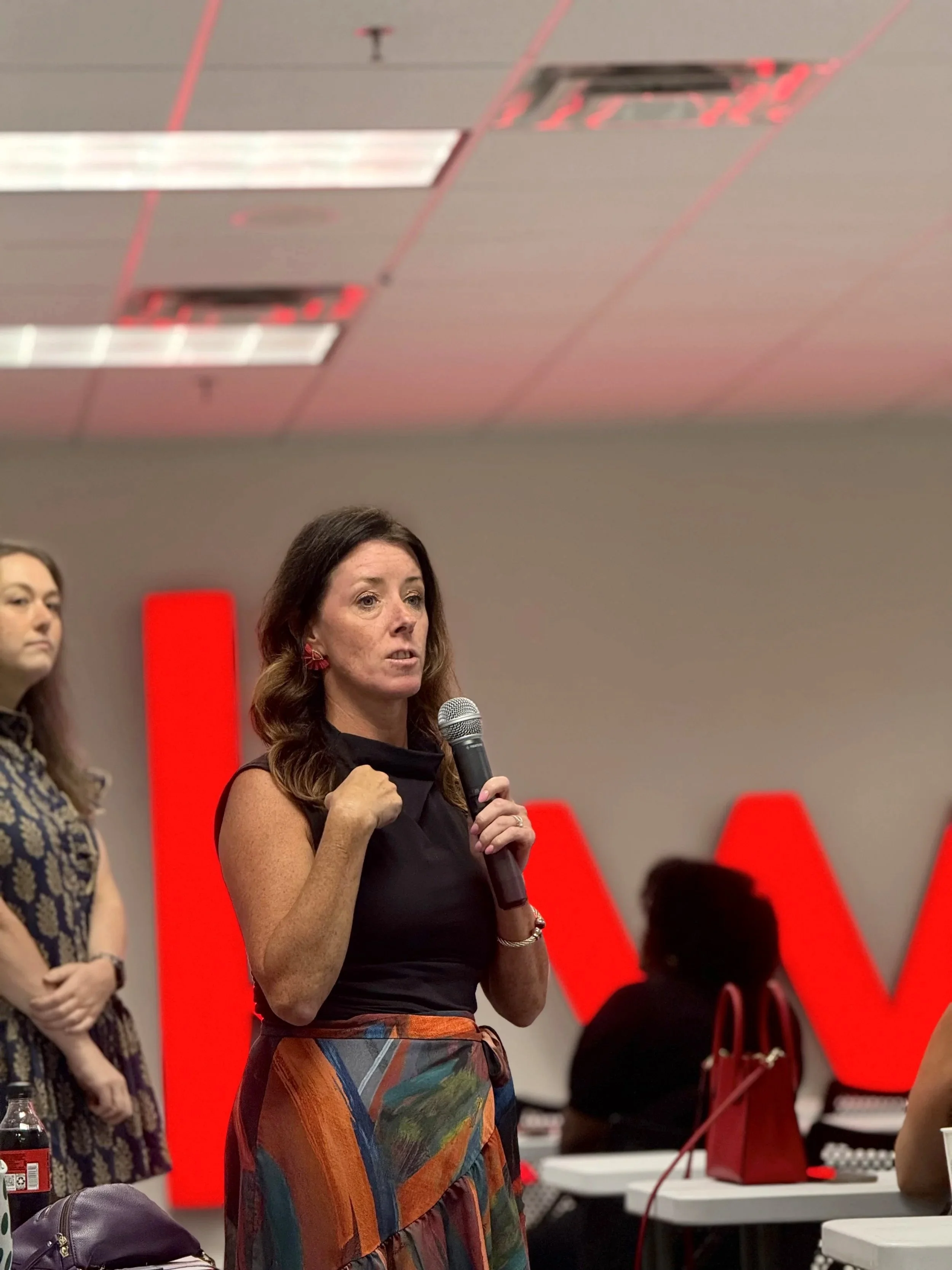 A woman with long wavy brown hair, wearing a sleeveless black top and a colorful patterned skirt, speaking into a microphone at an indoor event. In the background, there are large red letter blocks spelling 'W' and 'A', a woman with long dark hair and a patterned dress, and a table with a red bag, a water bottle, and other items.