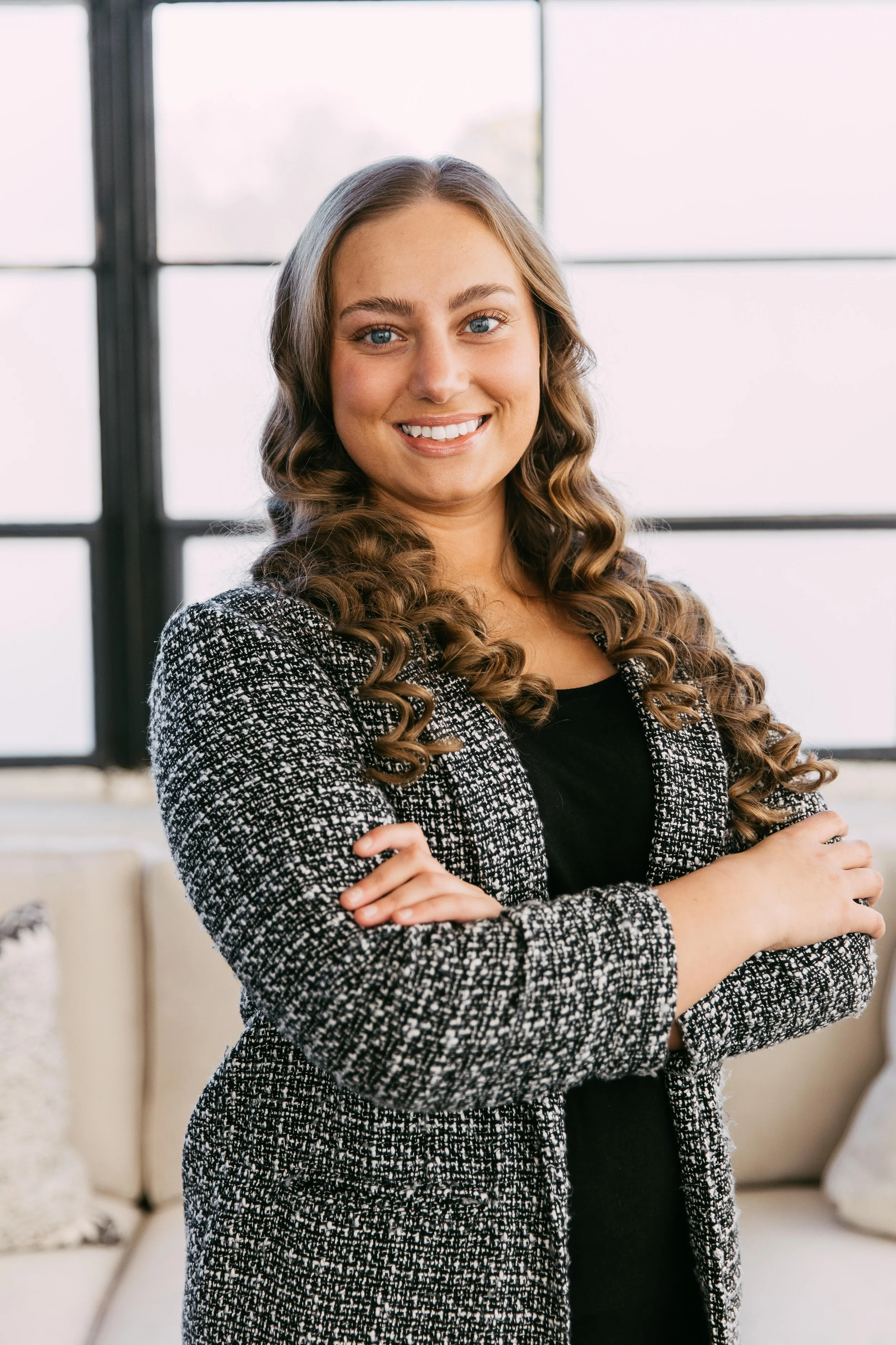 A woman with long, curly brown hair and blue eyes, smiling and wearing a black top and a black-and-white patterned blazer, standing in front of a large window in a modern office space.