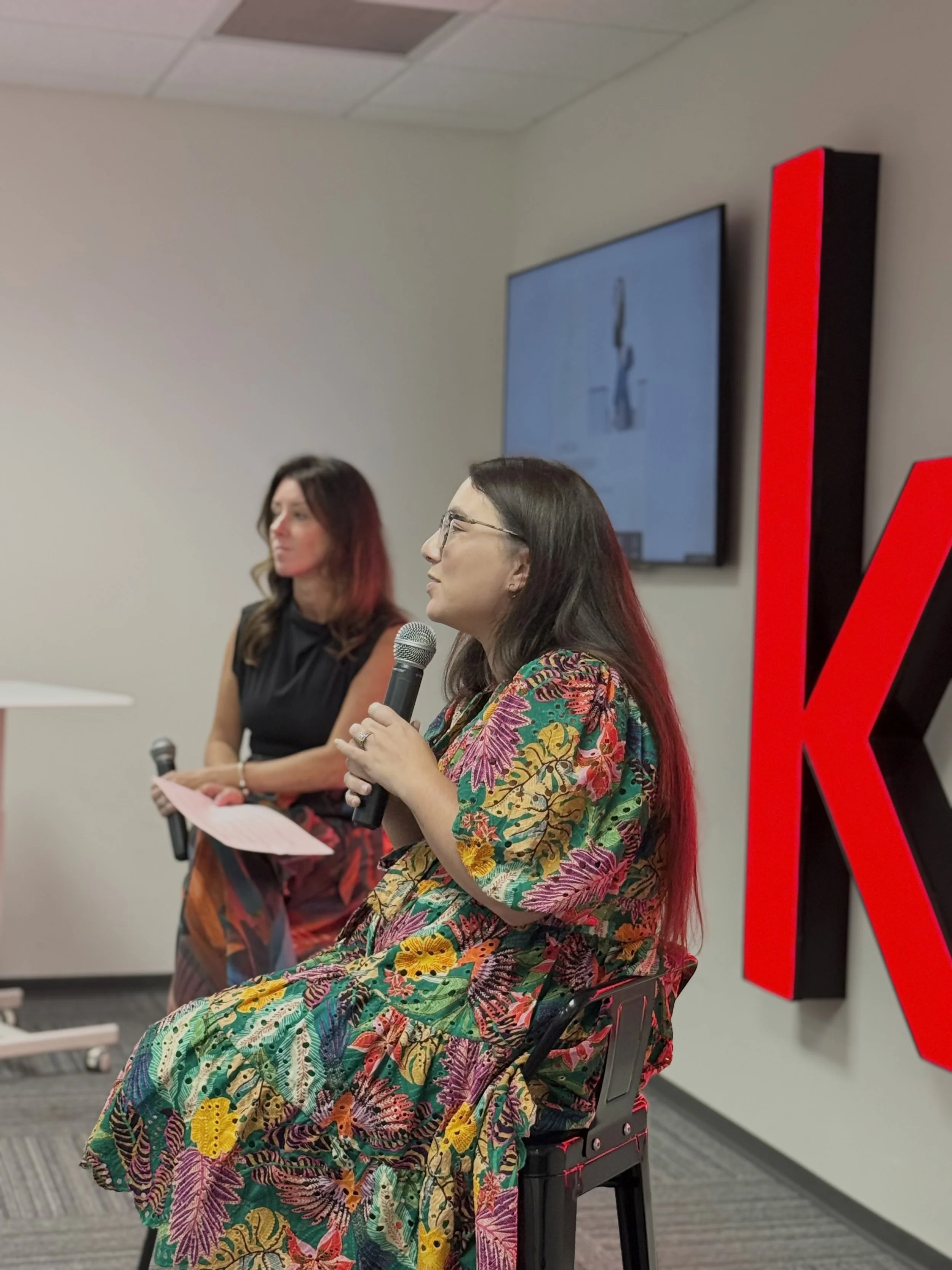 Two women sitting on chairs in a room, one is speaking into a microphone, and the other is holding a paper, with a large red and black letter V or K in the background, and a screen on the wall displaying some content.