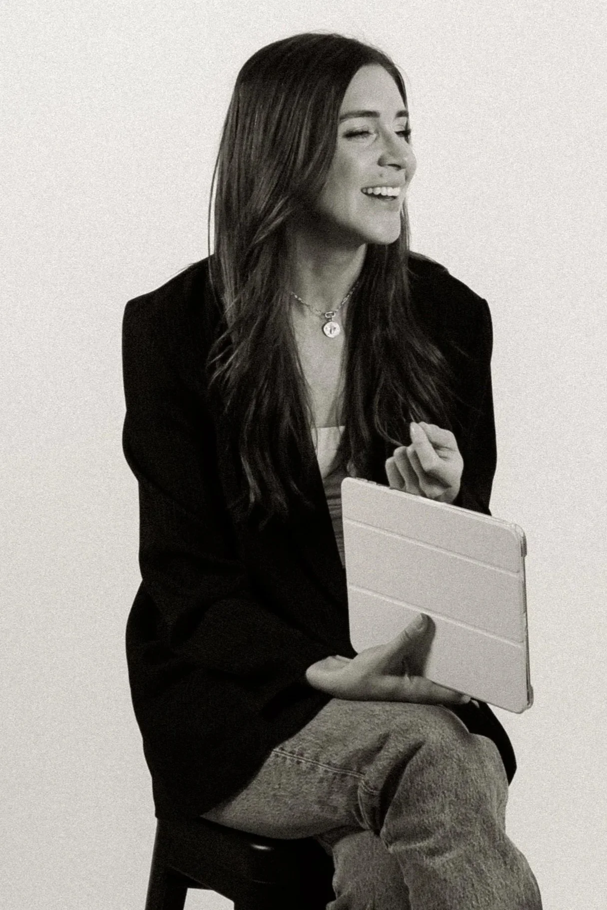A woman with long dark hair wearing a blazer, sitting on a chair, holding a tablet, and smiling with one eye closed against a plain background.