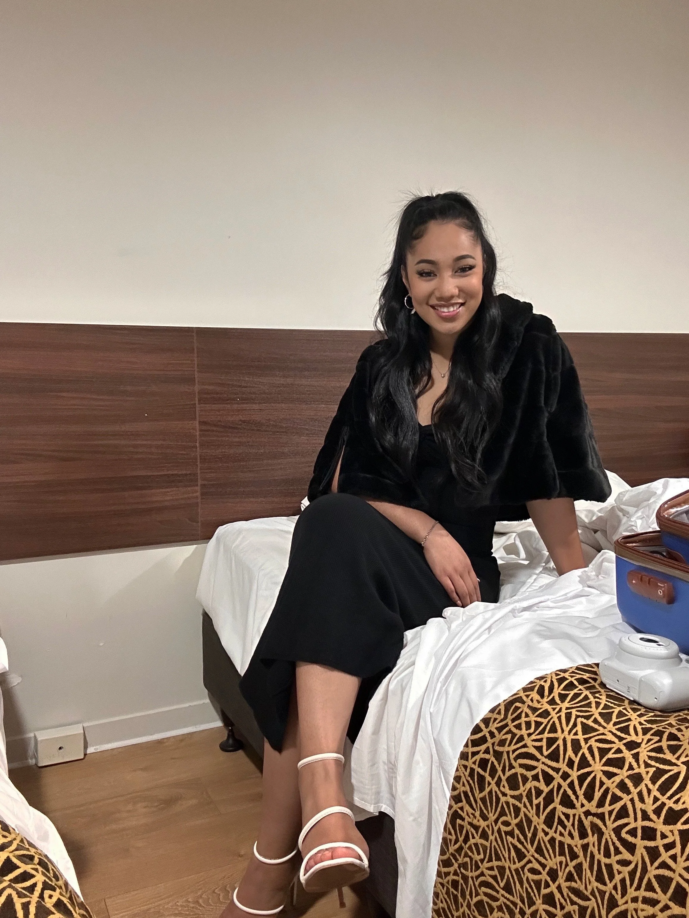 A smiling young woman sitting on a bed in an elegant black outfit.