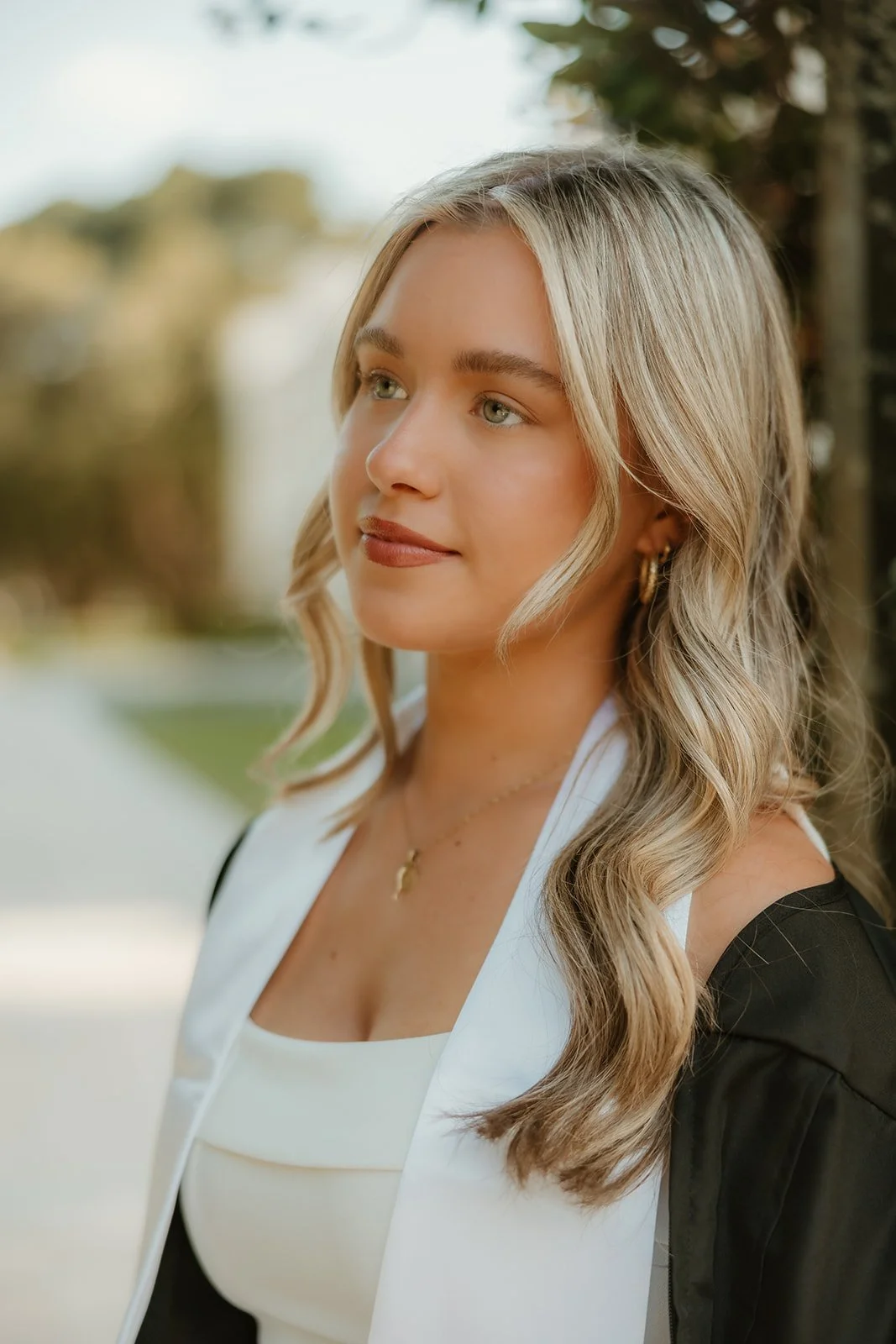 A young woman with blonde wavy hair, wearing a white top and a black graduation gown, standing outdoors with trees and a pathway in the background.
