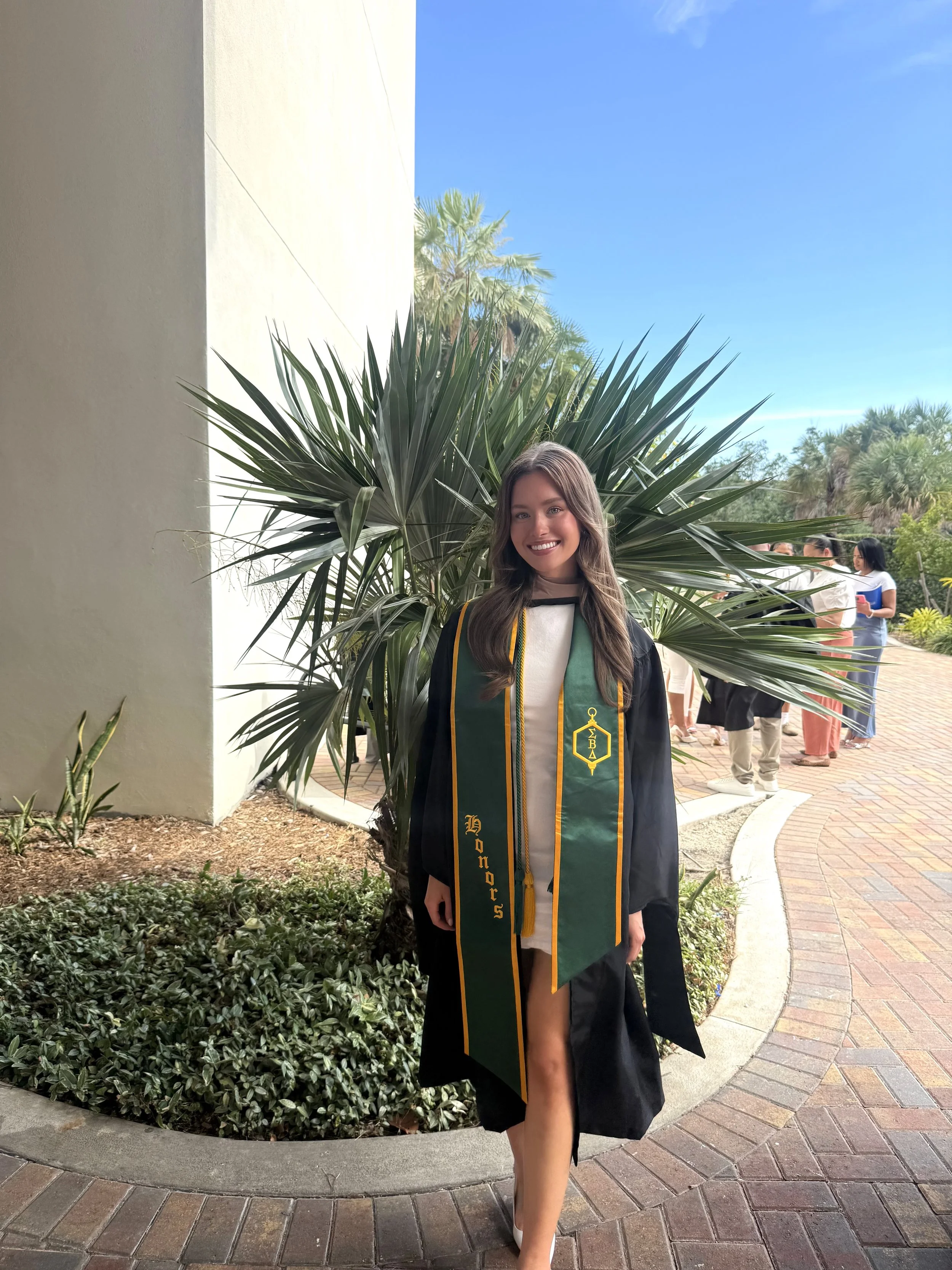 A young woman in a graduation cap and gown standing outdoors in front of a large green plant, smiling. She is wearing a green stole with gold lettering and a black gown, and the background shows a group of people and clear blue sky.