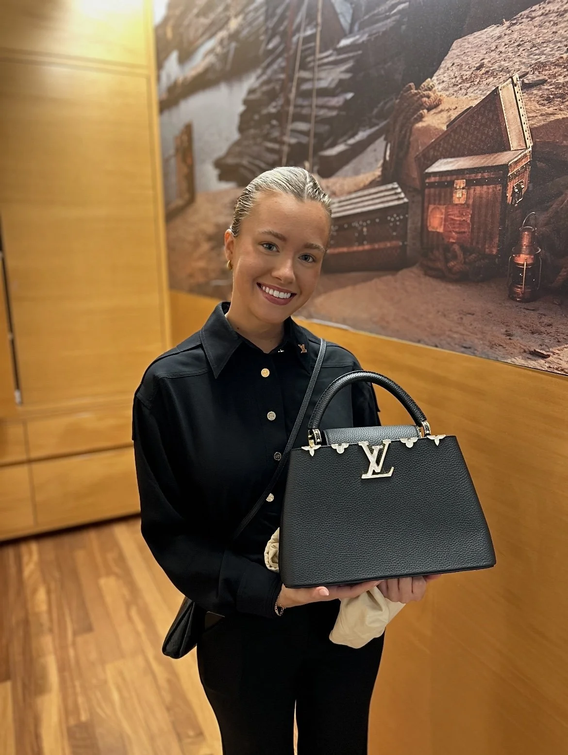 A smiling young woman holding a black Louis Vuitton handbag with gold hardware, standing in a room with wooden floors and a wall decorated with a mountain landscape mural.