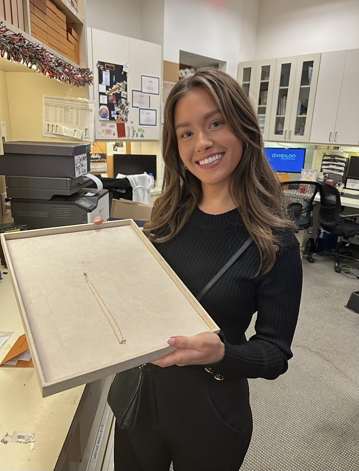 A young woman with long brown hair wearing a black ribbed sweater holding a jewelry display box with a gold necklace inside, smiling in an office setting.