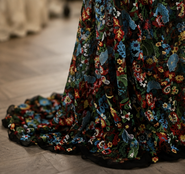 Close-up of a wedding dress with floral embroidery on a black fabric, train lying on a wooden floor.