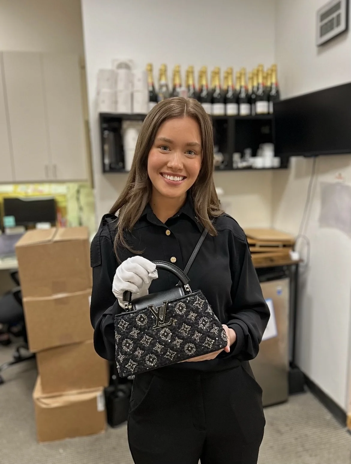 A smiling woman in black clothing holding a designer handbag in an indoor setting, with bottles on a shelf in the background.