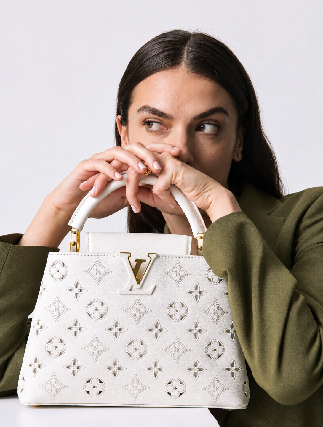 A woman with dark hair, wearing a green jacket, holding a white designer handbag with gold accents and open pattern, partially covering her face while looking to the side.