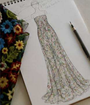 Fashion design sketch of a woman in a floral, floor-length dress with detailed patterns, placed on a white surface next to a black pen and a colorful embroidered fabric.