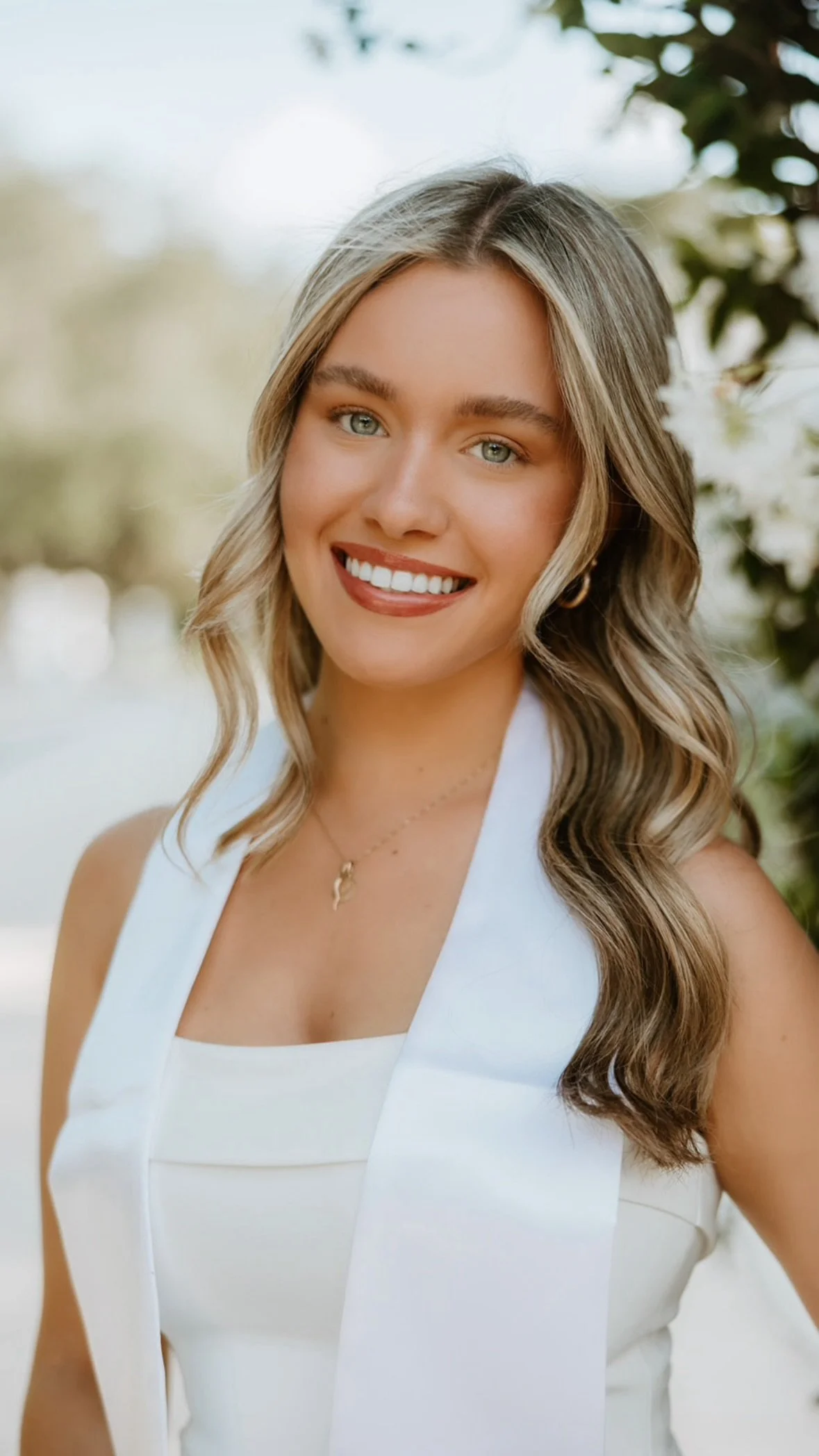 A young woman with long wavy blonde hair, blue eyes, and a bright smile, wearing a white sleeveless dress and a delicate necklace, outdoors with blurred greenery in the background.