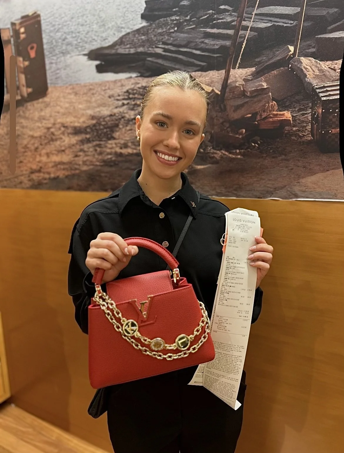 A smiling woman with blonde hair pulled back, holding a red luxury handbag with chain detail and a long receipt. She is standing inside a store with a wooden wall and a nature-themed background mural.