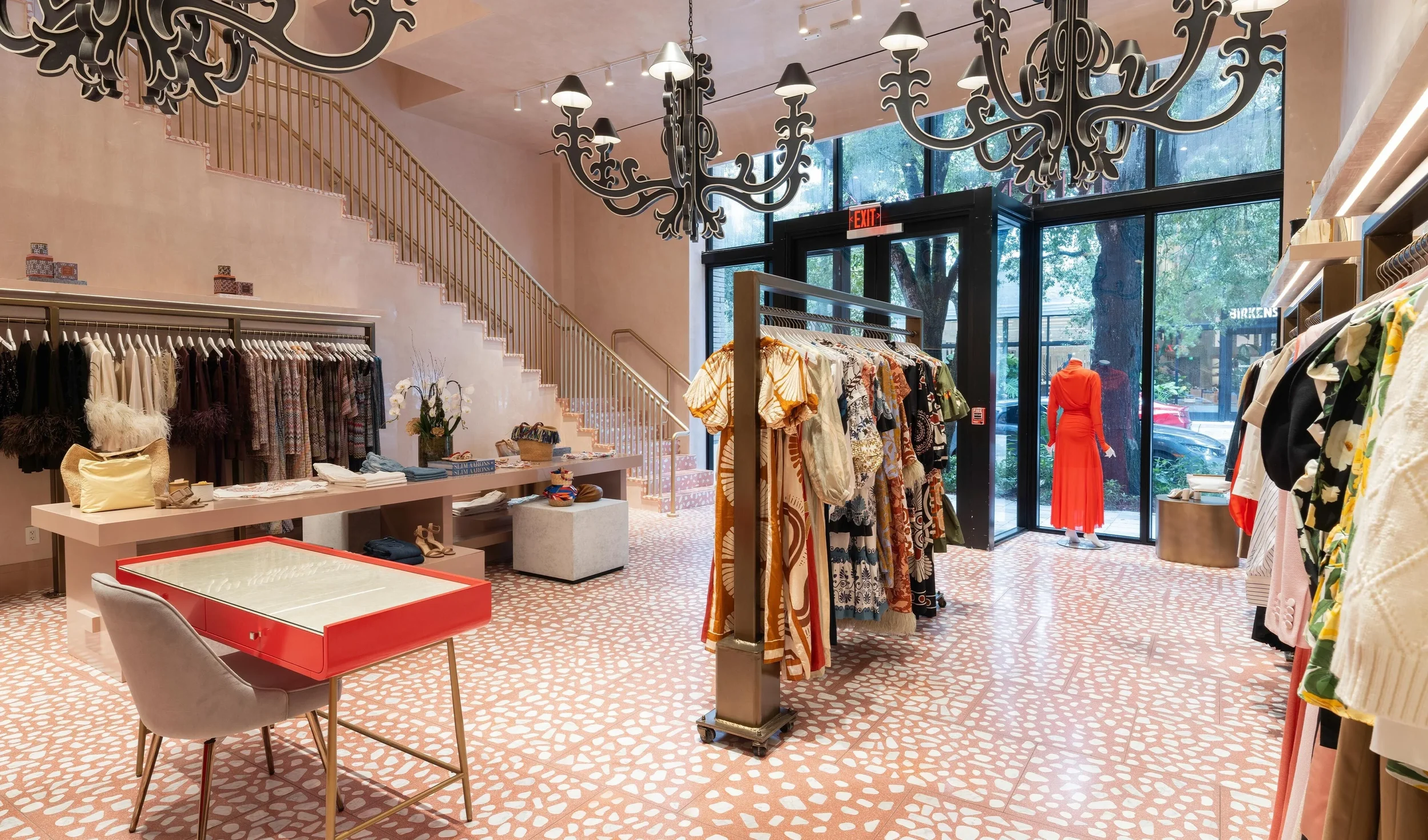 A boutique clothing store with clothing racks, a display table, and a staircase. Large windows at the front let in natural light, with mannequins dressed in orange and yellow clothing visible outside.