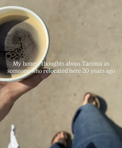 A person holding a cup of black coffee taken from a top-down view, with a blurry background showing the person’s legs and feet on a sandy surface, and text overlay that says, "My honest thoughts about Tacoma as someone who relocated here 20 years ago."