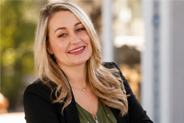 Smiling woman with blonde hair, wearing a black blazer over a green top, outdoors with blurred trees in the background.