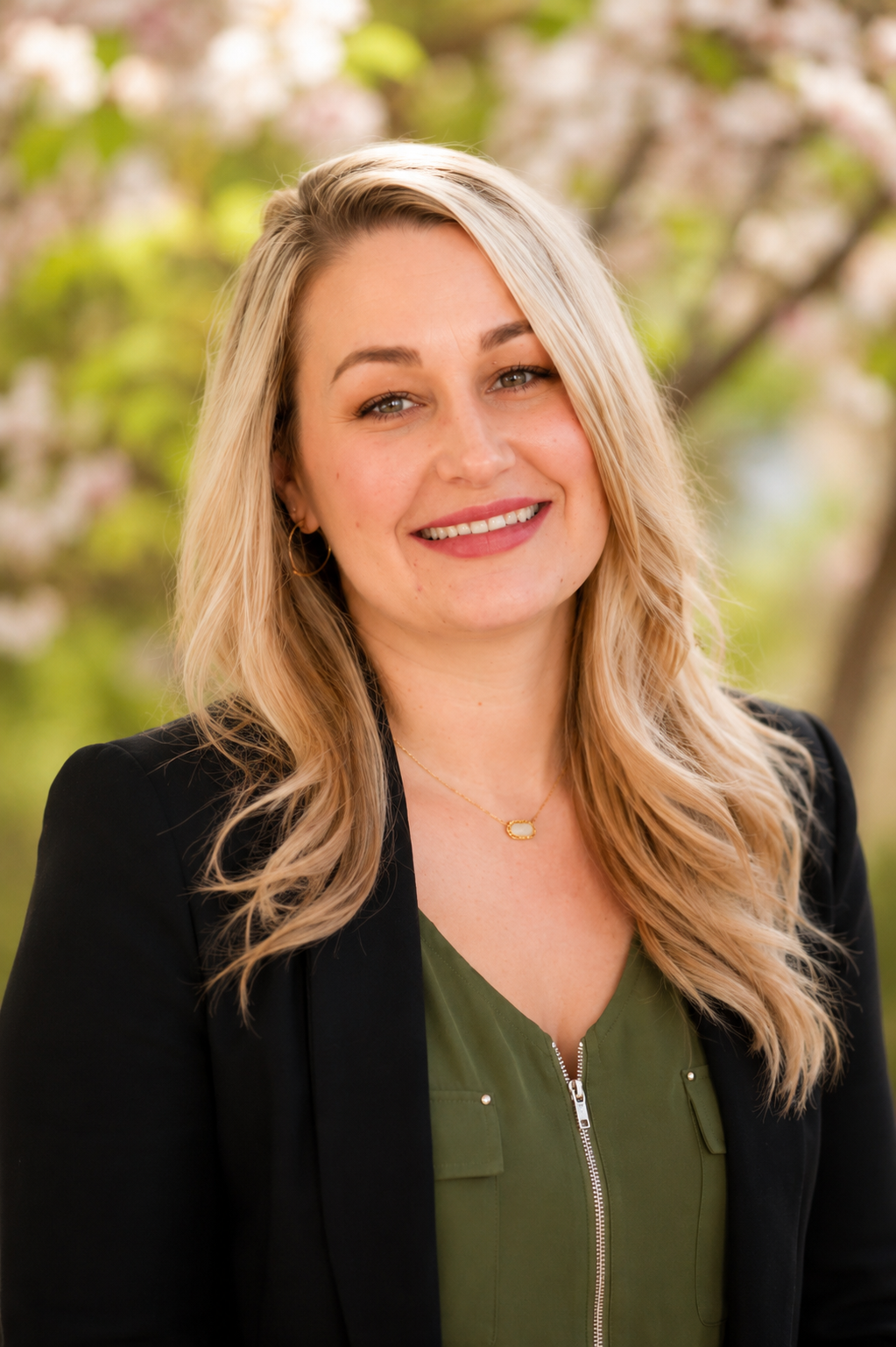 A woman with blonde, wavy hair smiling outdoors with blooming trees in the background, wearing a black blazer and green top.