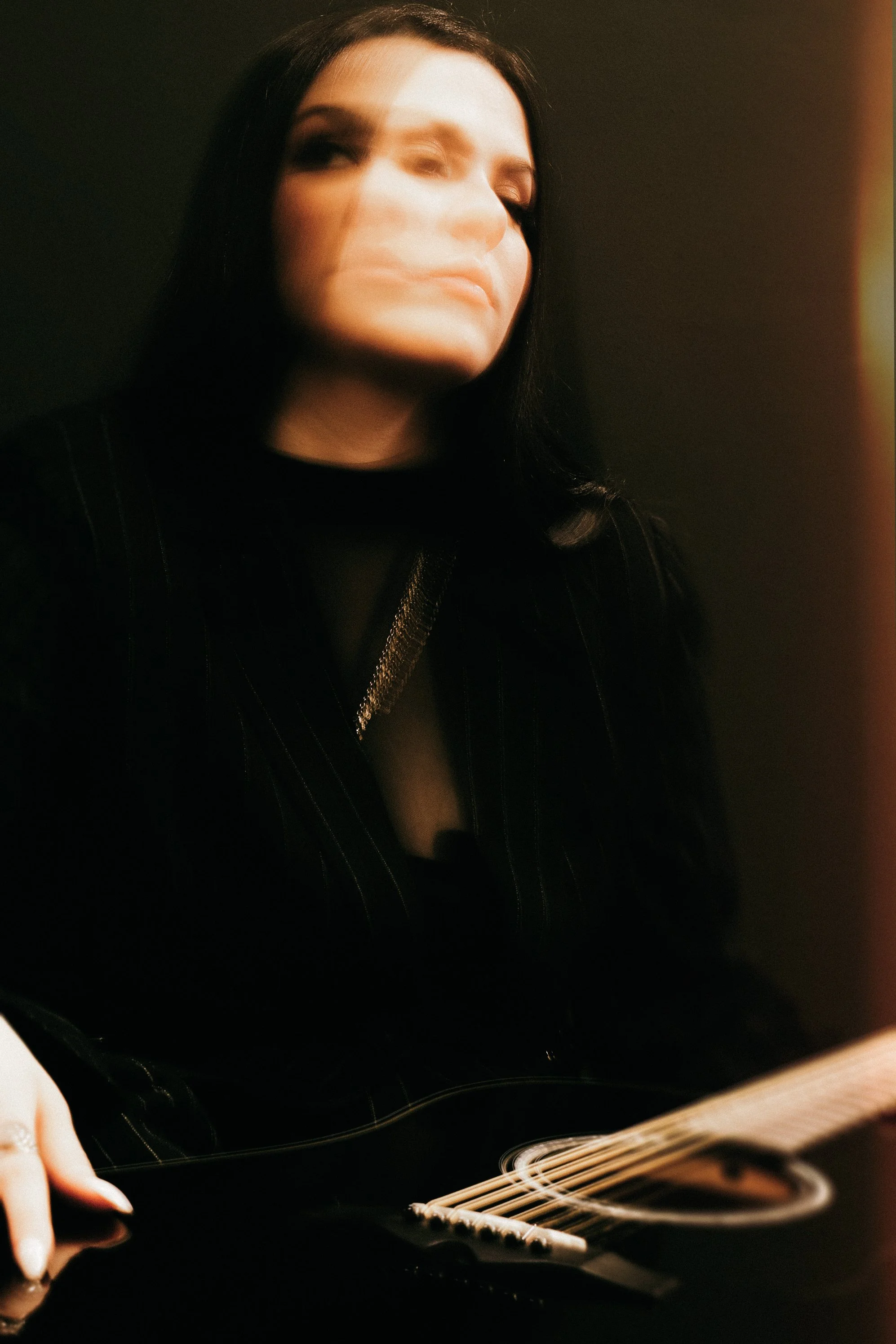 A woman with dark hair and a black striped suit playing a black acoustic guitar with a blurred face.