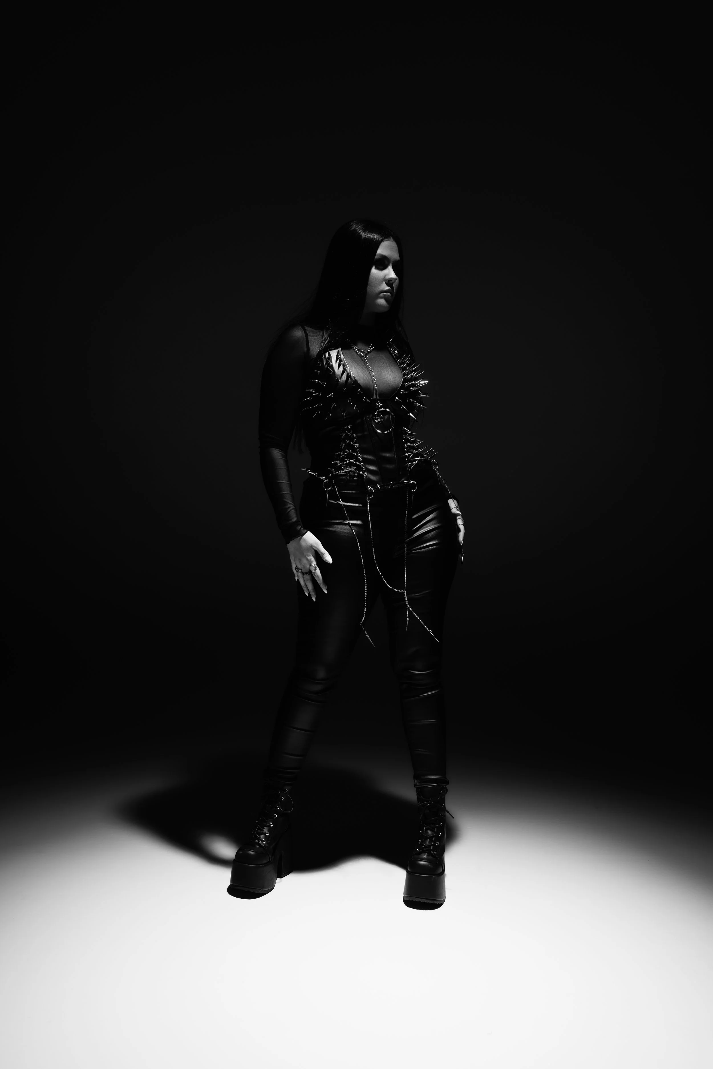 A woman with dark hair standing in a spotlight with a dark background, wearing a leather outfit adorned with metal spikes and chains, with platform boots.